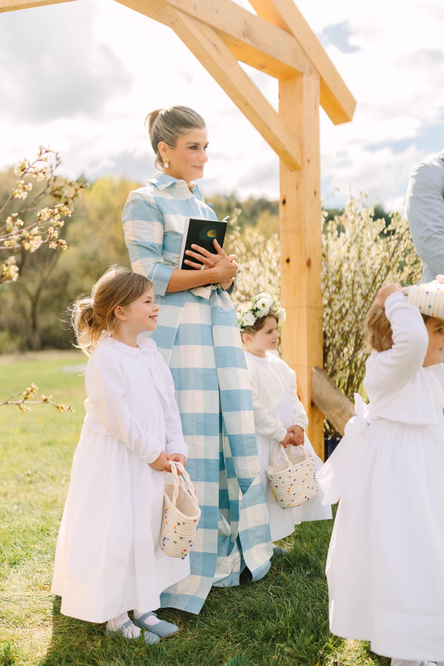 Officiant in blue striped gown holds bible as flower girls in white dresses line up at outdoor altar.