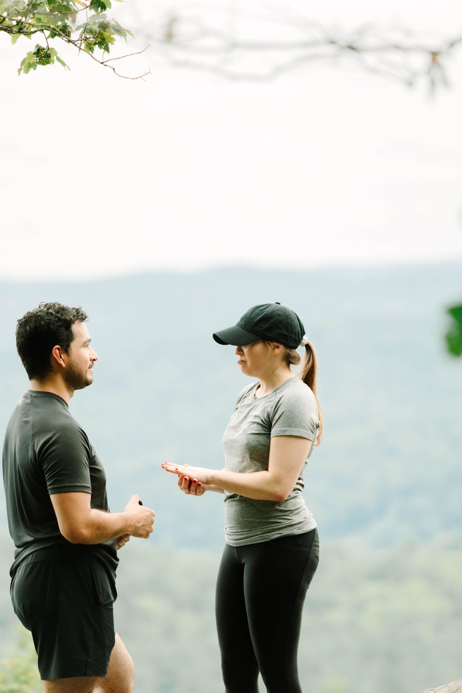 Two athletes in gray workout clothes discuss their run at a scenic mountain overlook surrounded by lush green trees.