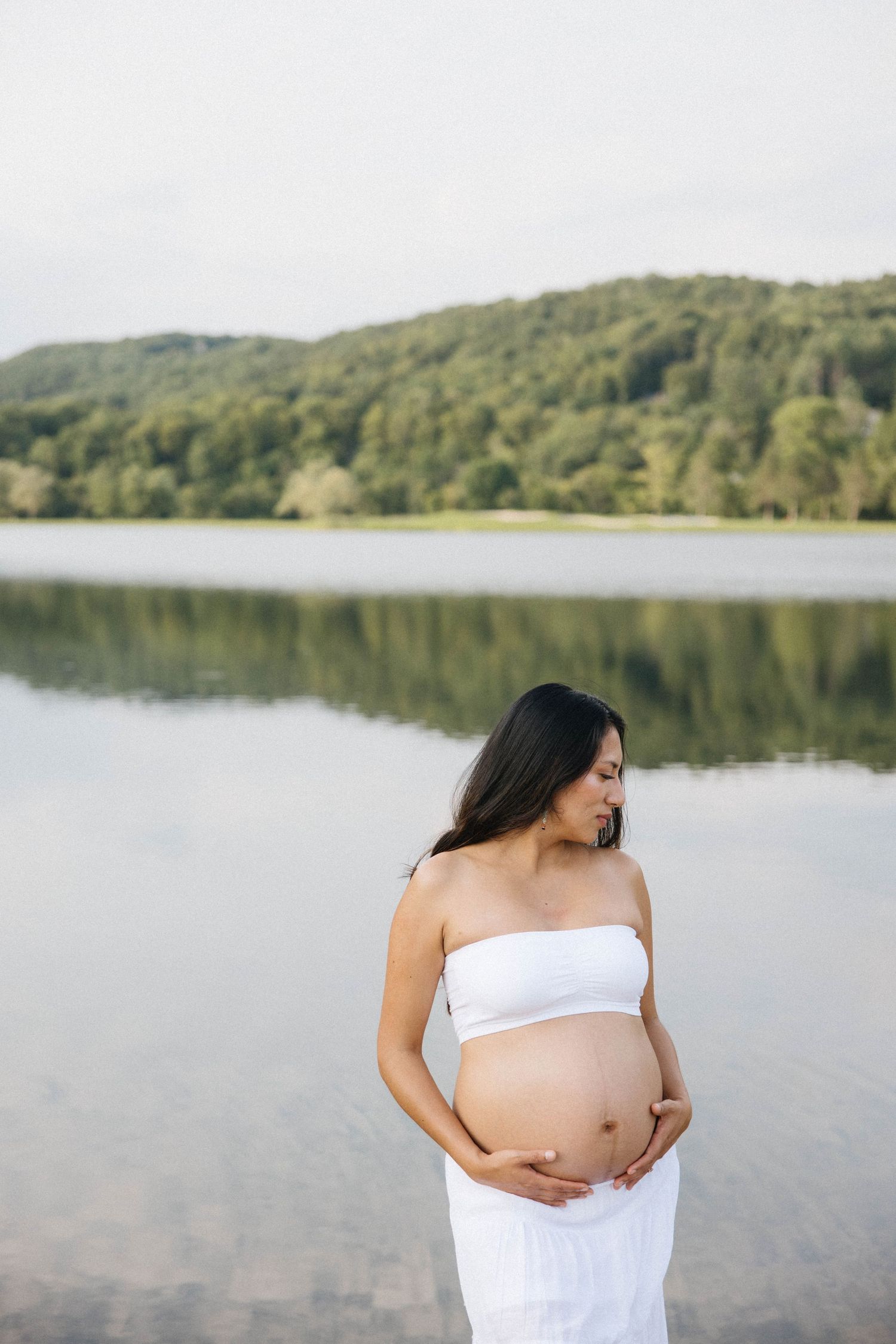 Pregnant woman in white dress cradling her baby bump by a serene lake with lush green hills reflected in the water.