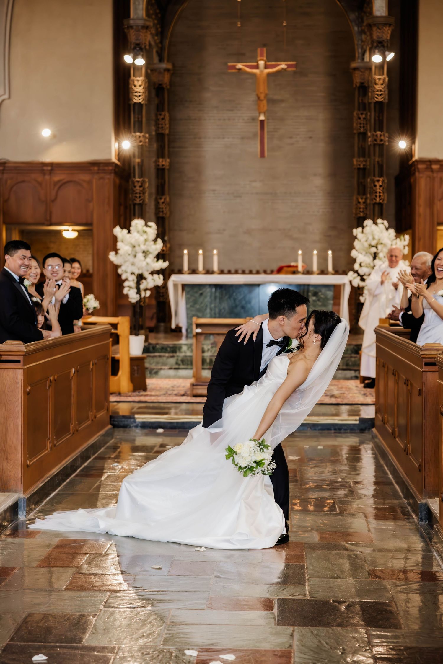 Groom dipping bride for a kiss at church altar while wedding guests applaud in background.