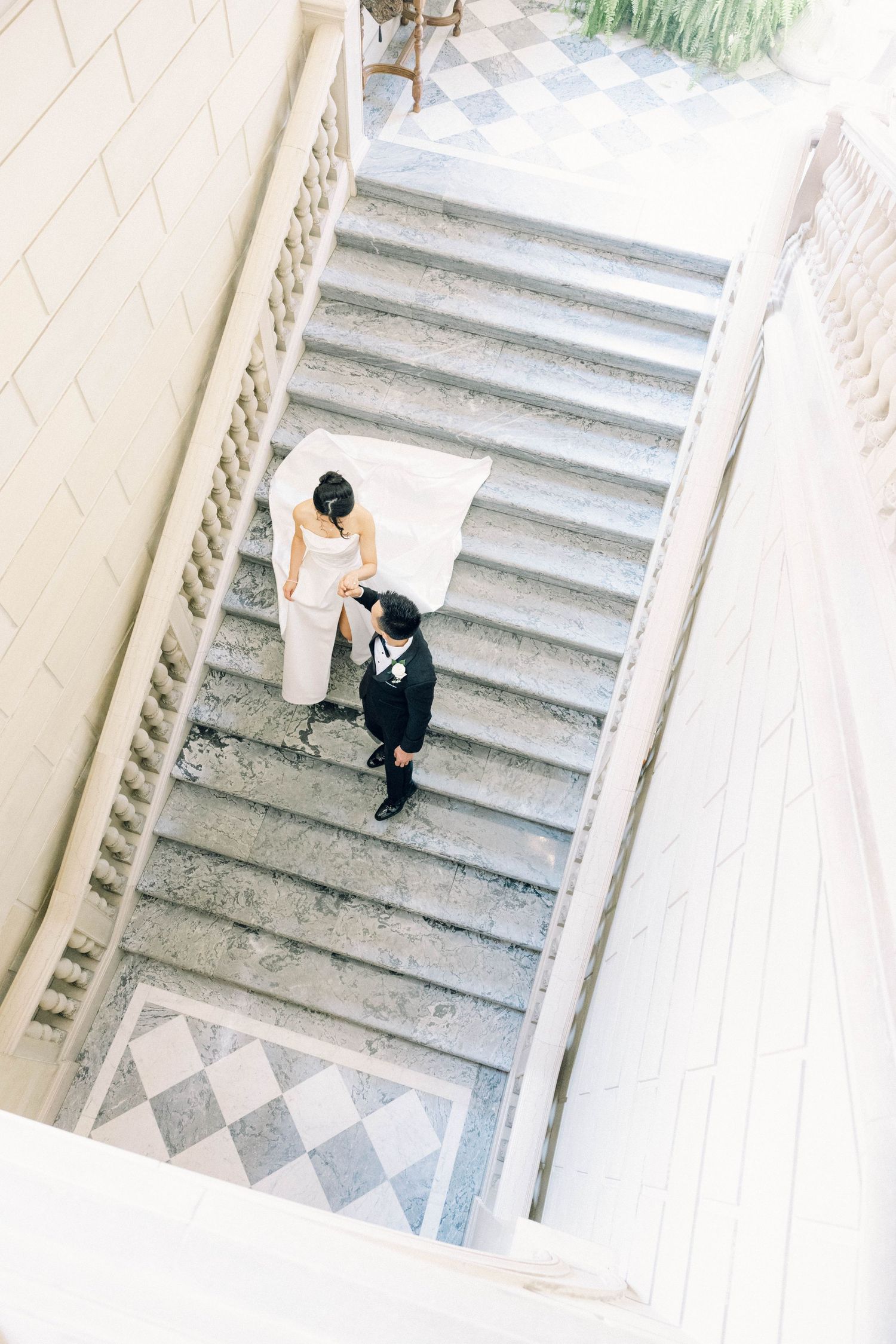 Aerial view of bride and groom holding hands while walking up marble staircase together.