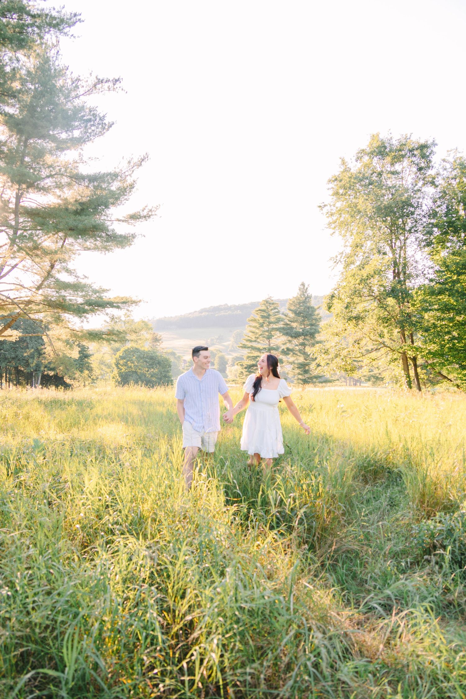 Couple holding hands walking through a sunlit meadow with lush green grass and trees during golden hour.