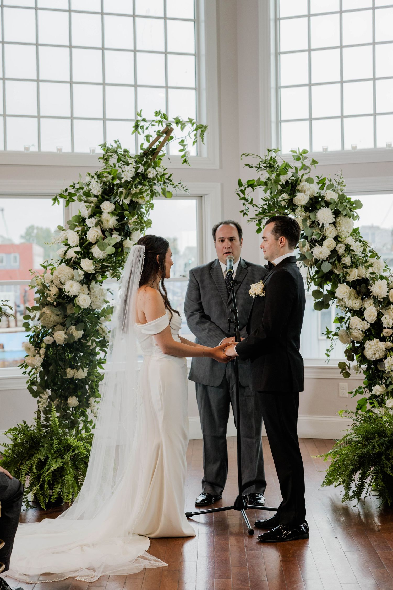 Bride and groom exchange vows under a lush floral arch with white roses and greenery during an indoor wedding ceremony.