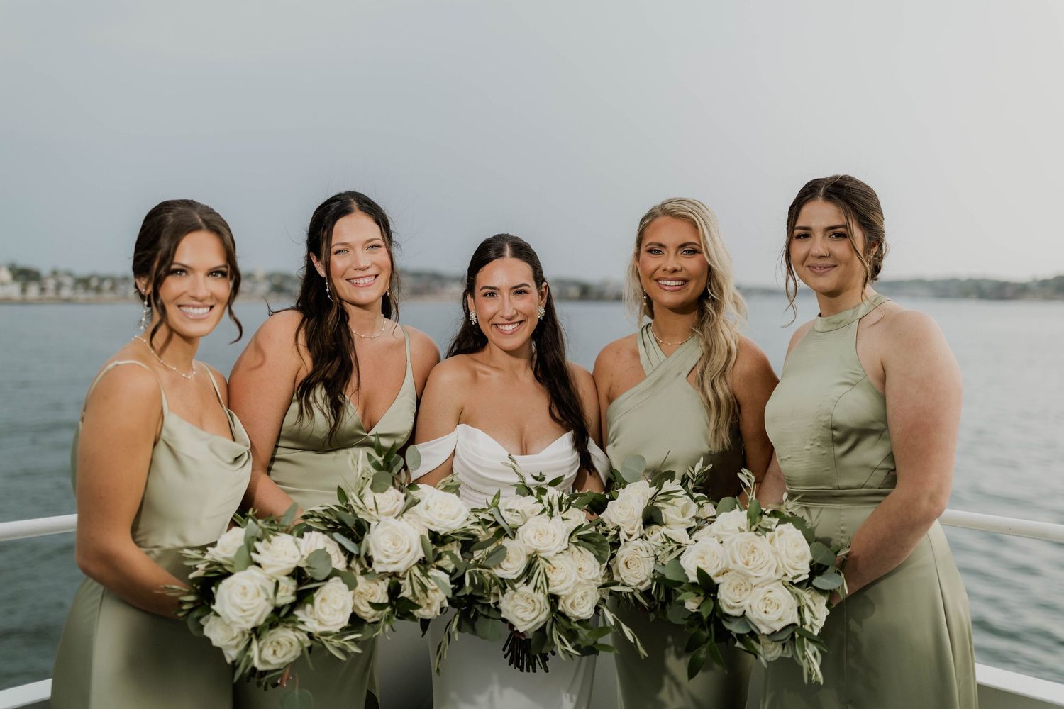 Bride poses with four bridesmaids in sage green gowns holding white rose bouquets on waterfront.