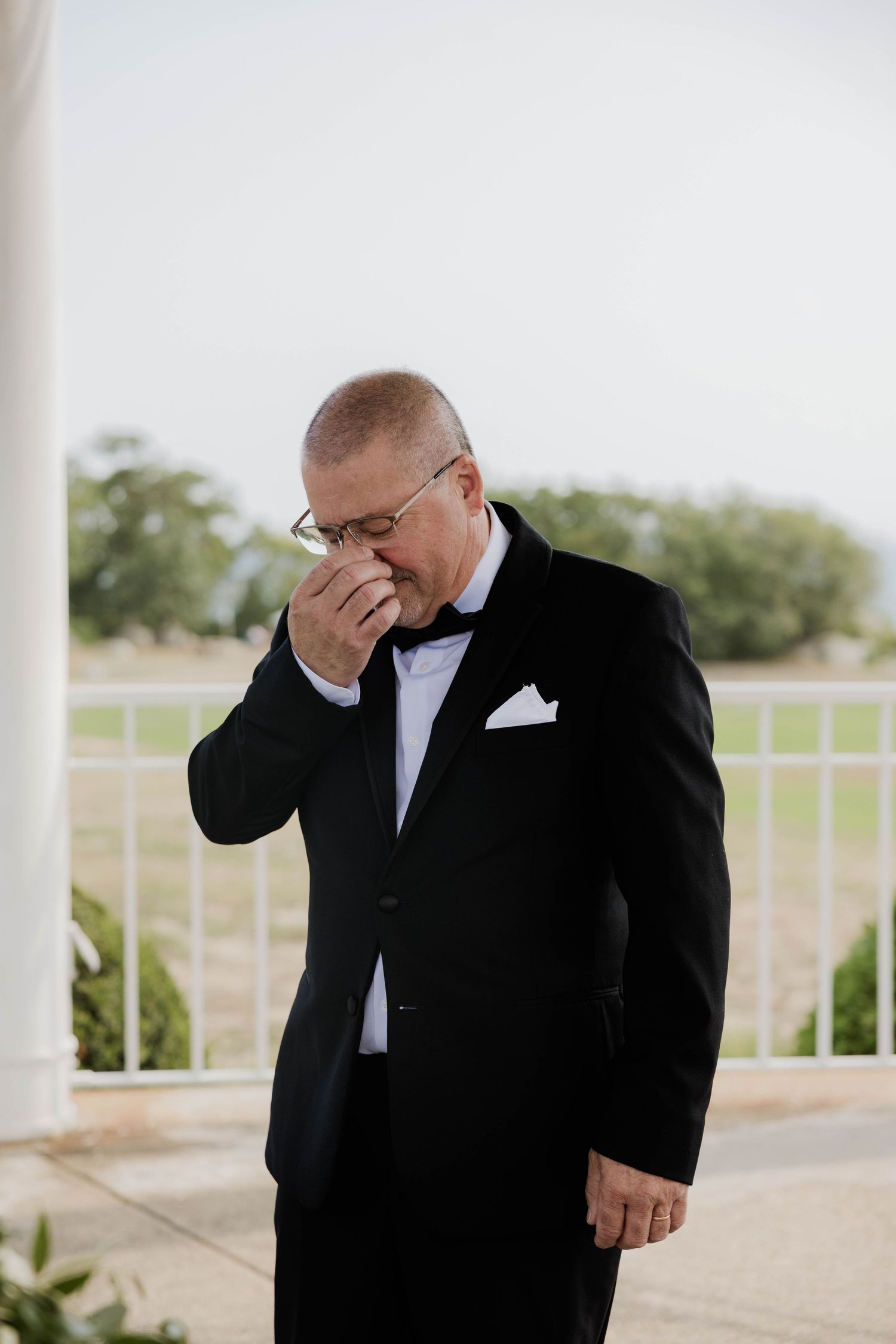 Emotional groom in black tuxedo wiping tears before wedding ceremony outdoors.