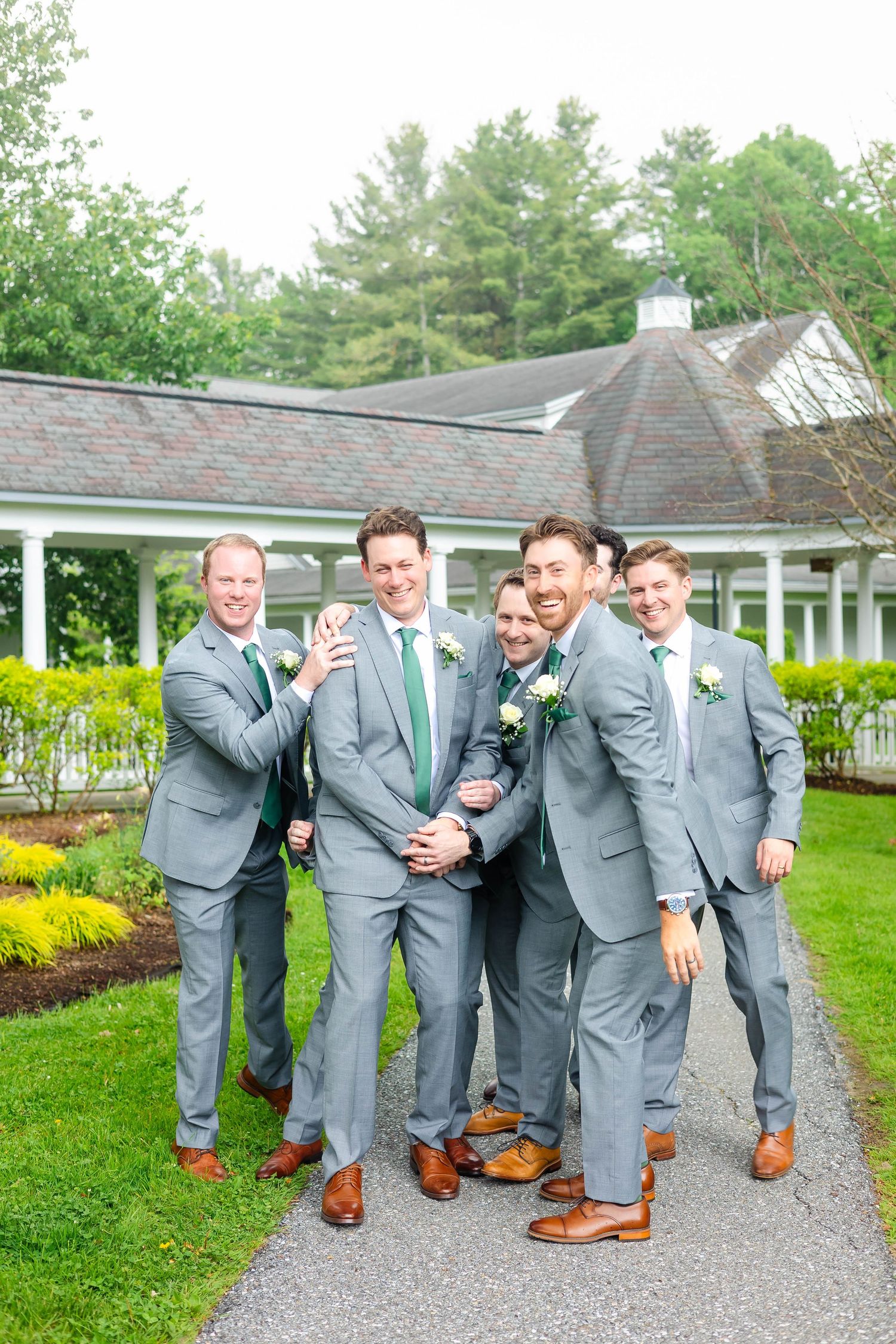 Groomsmen in gray suits and green ties laugh and pose together on garden pathway.