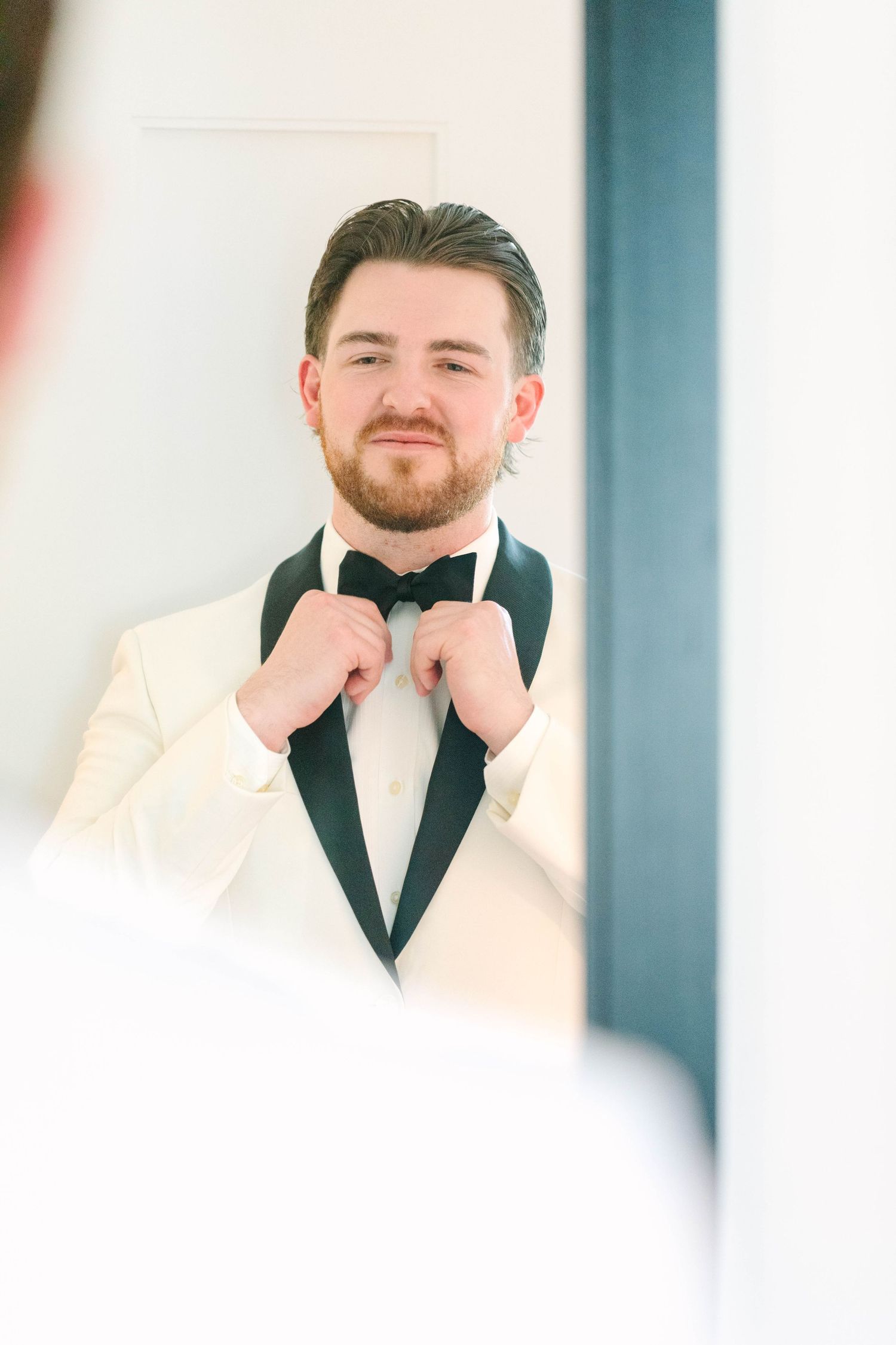 Dapper groom in a white tuxedo jacket adjusts his black bow tie while looking in a mirror.