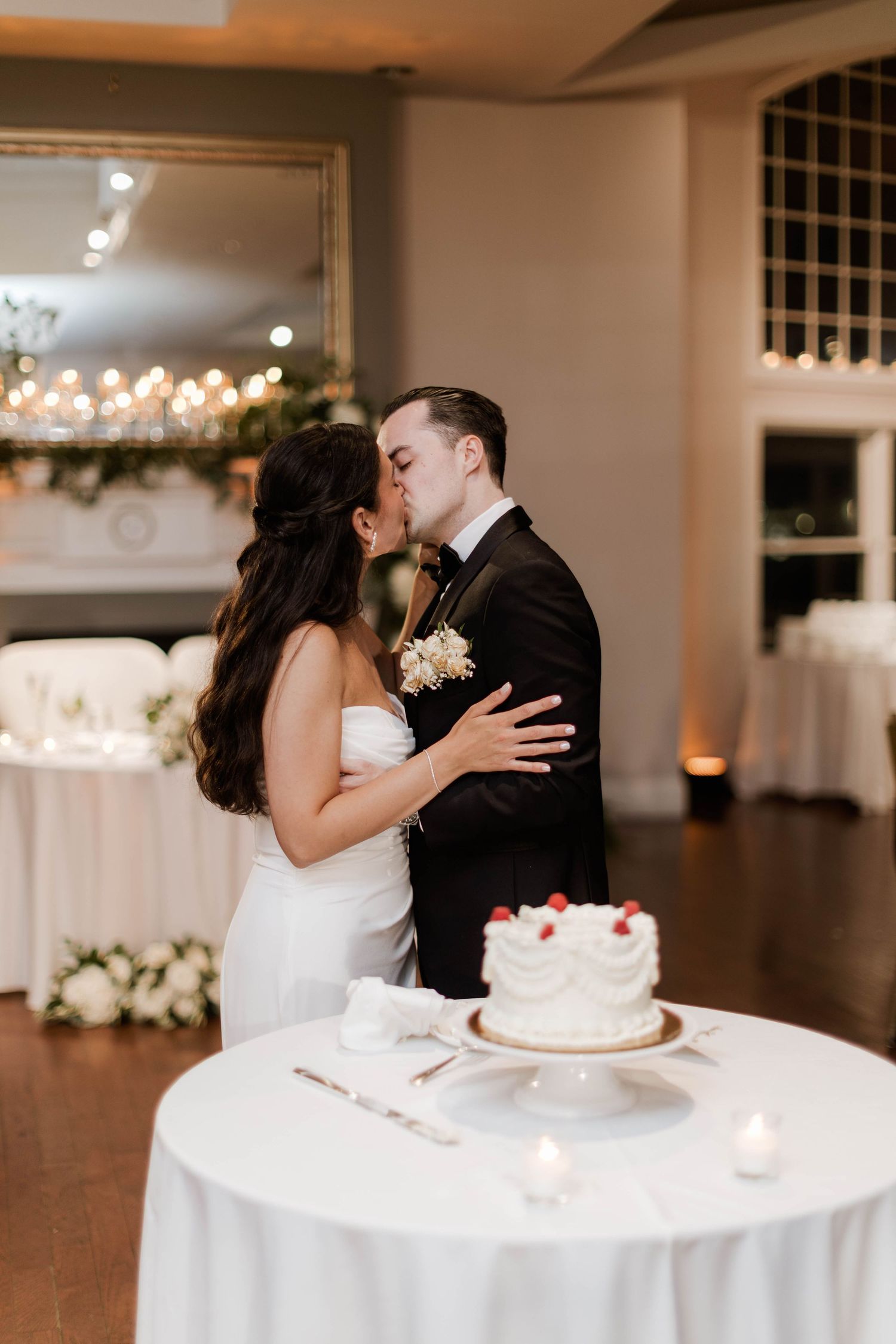 Bride and groom share romantic kiss after cutting white strawberry wedding cake at reception.