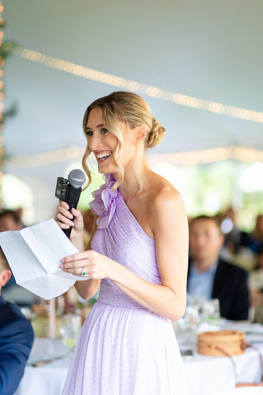 Bridesmaid in lavender one-shoulder gown delivering heartfelt speech with microphone at outdoor wedding reception.