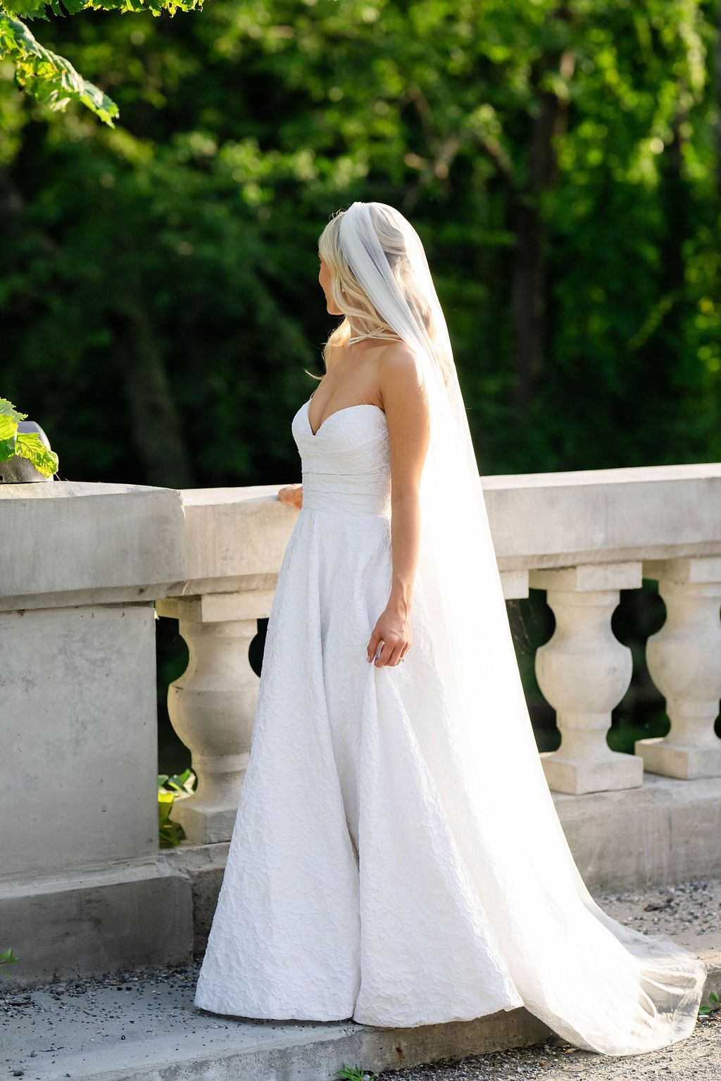 Elegant bride in strapless white lace gown and cathedral veil stands by stone balustrade outdoors.
