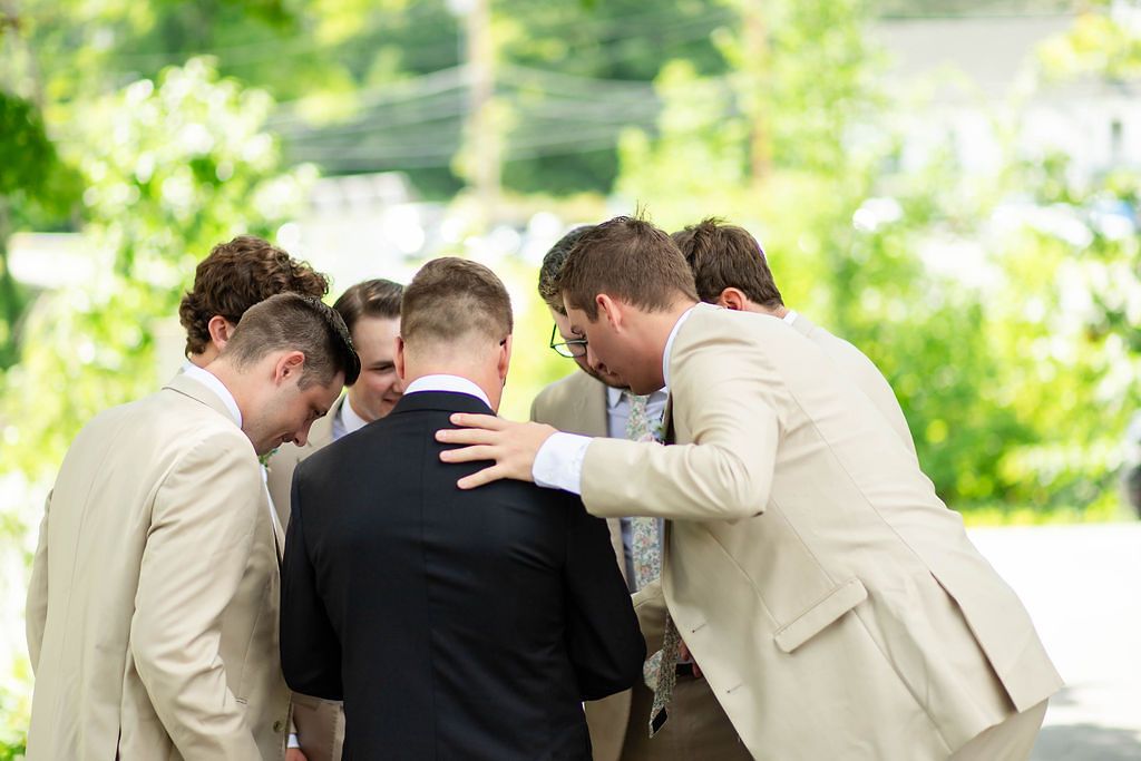 Groomsmen in tan suits huddle together outdoors in a circle of prayer before the wedding.