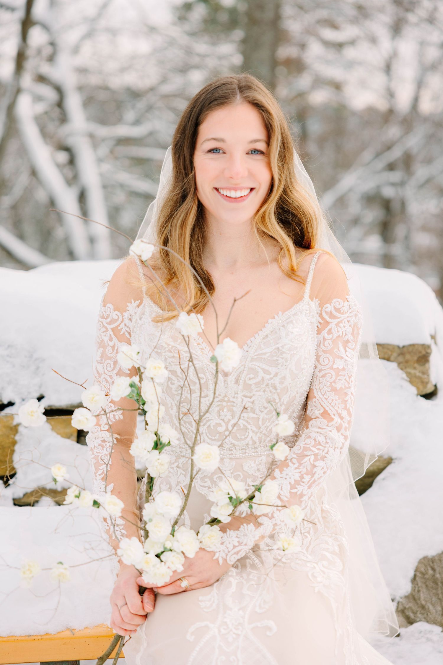 Smiling bride in lace wedding dress with veil holding white floral bouquet in winter snow.