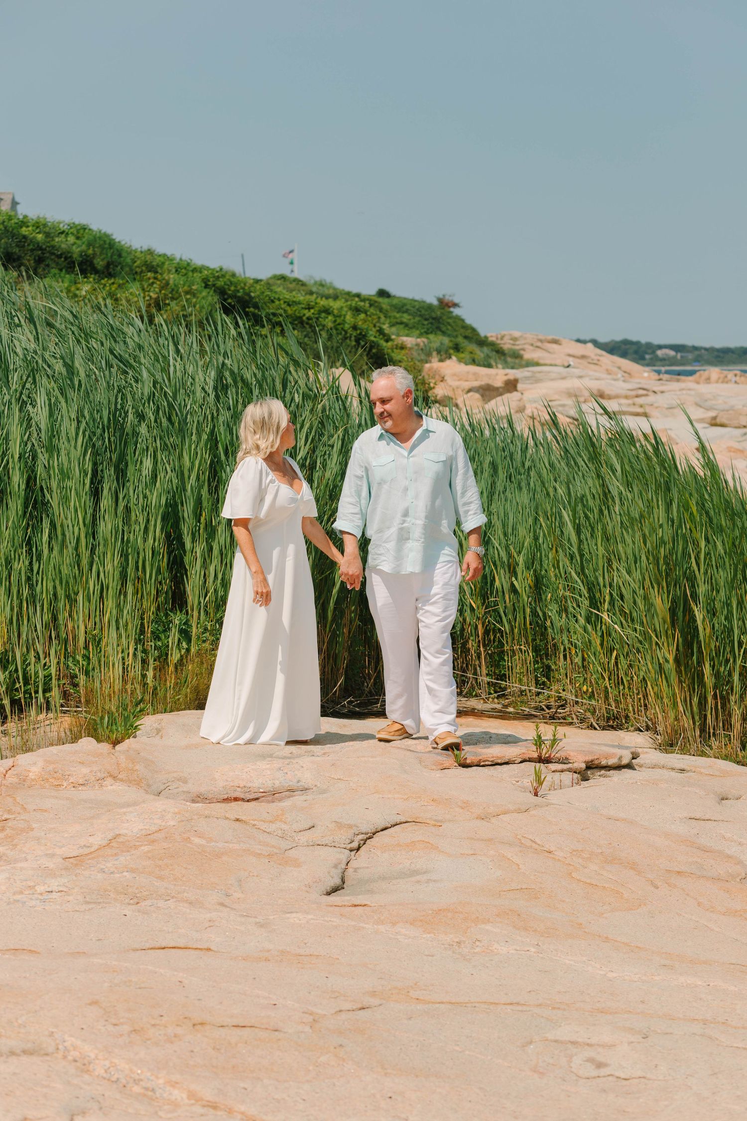 Older couple in white outfits holding hands on rocky shore with tall green reeds behind them.