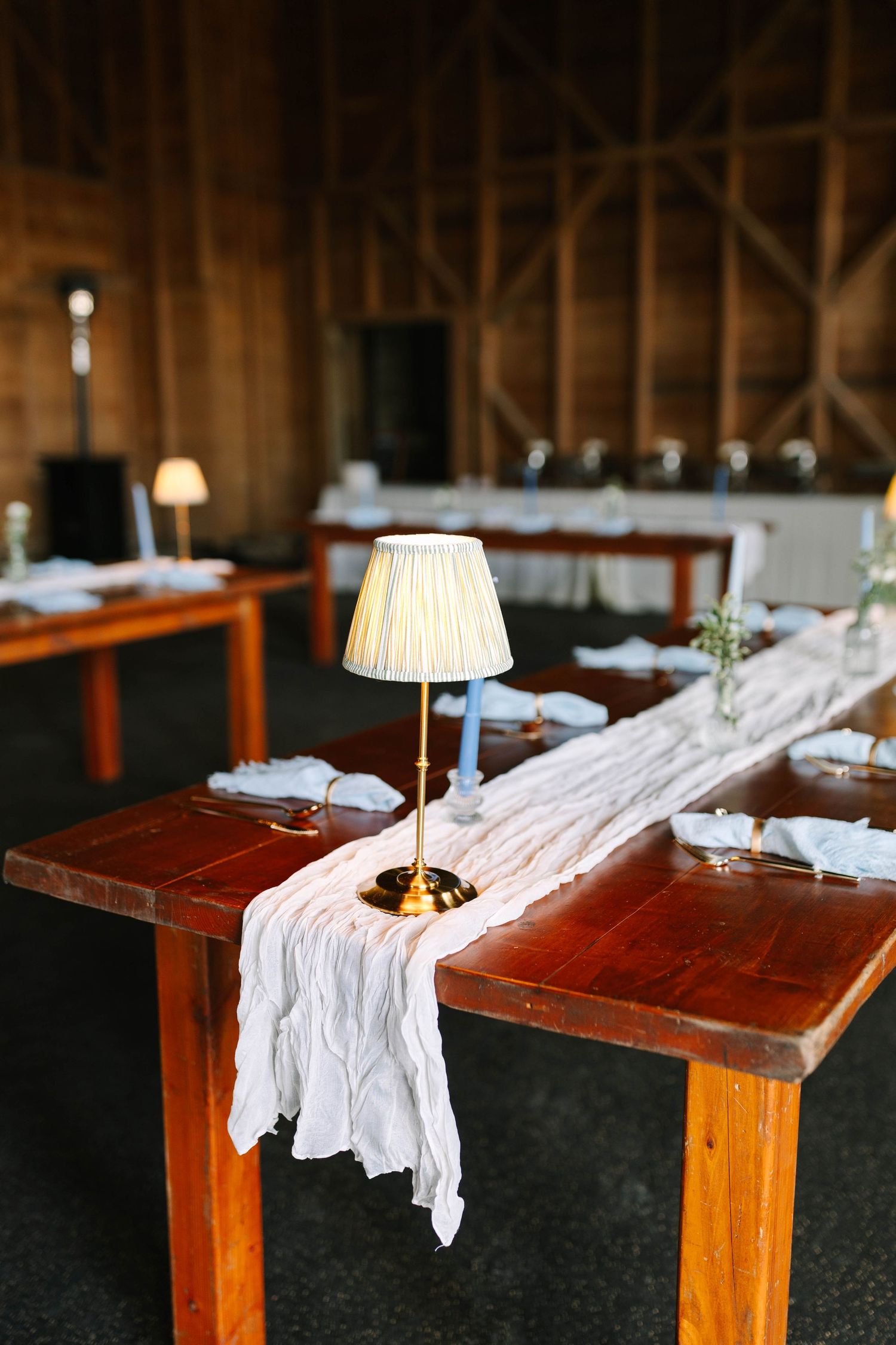 Rustic barn wedding reception tables with white linen runners and gold table lamps.