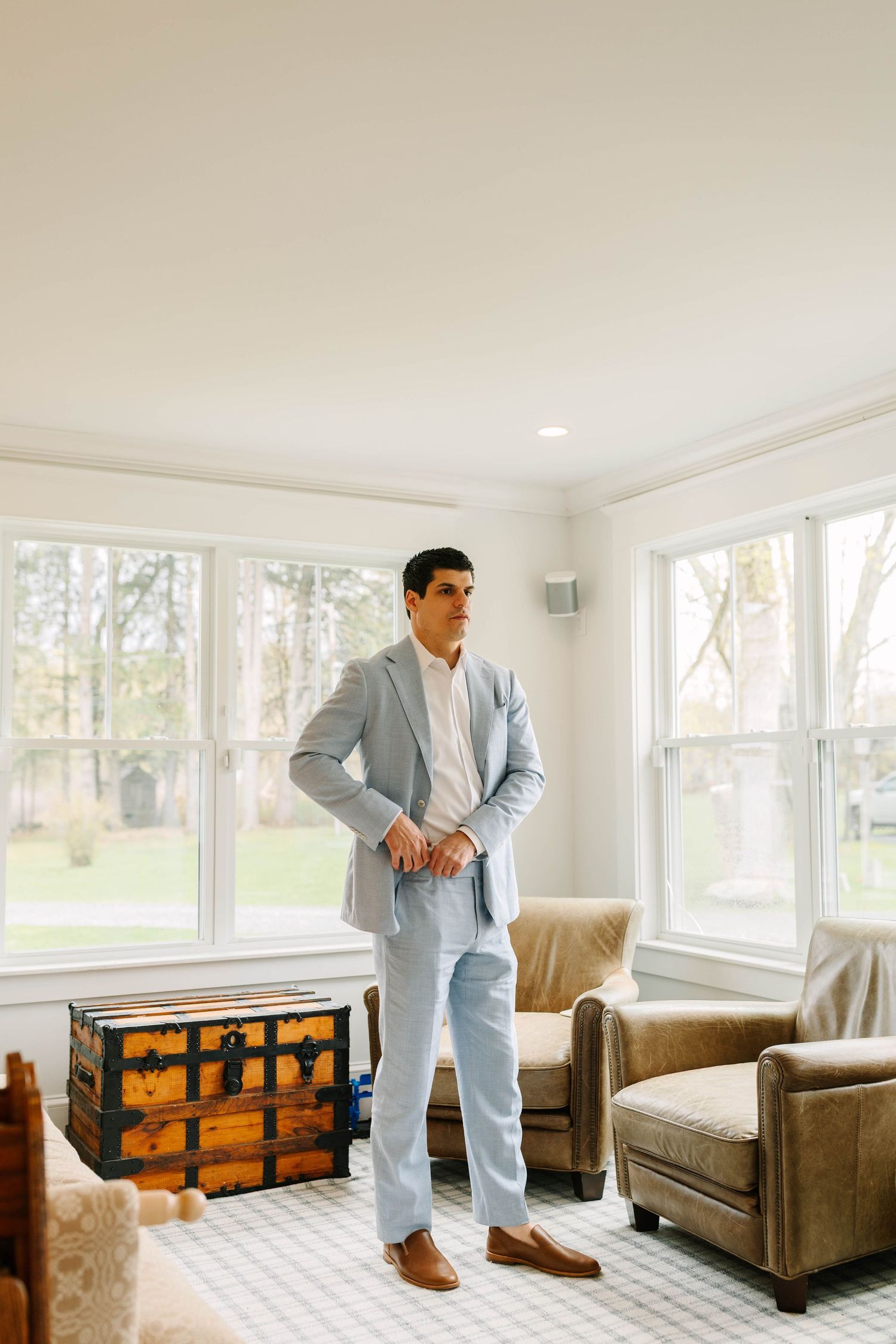 Groom in a light blue suit standing in a sunlit room with large windows and vintage trunk.
