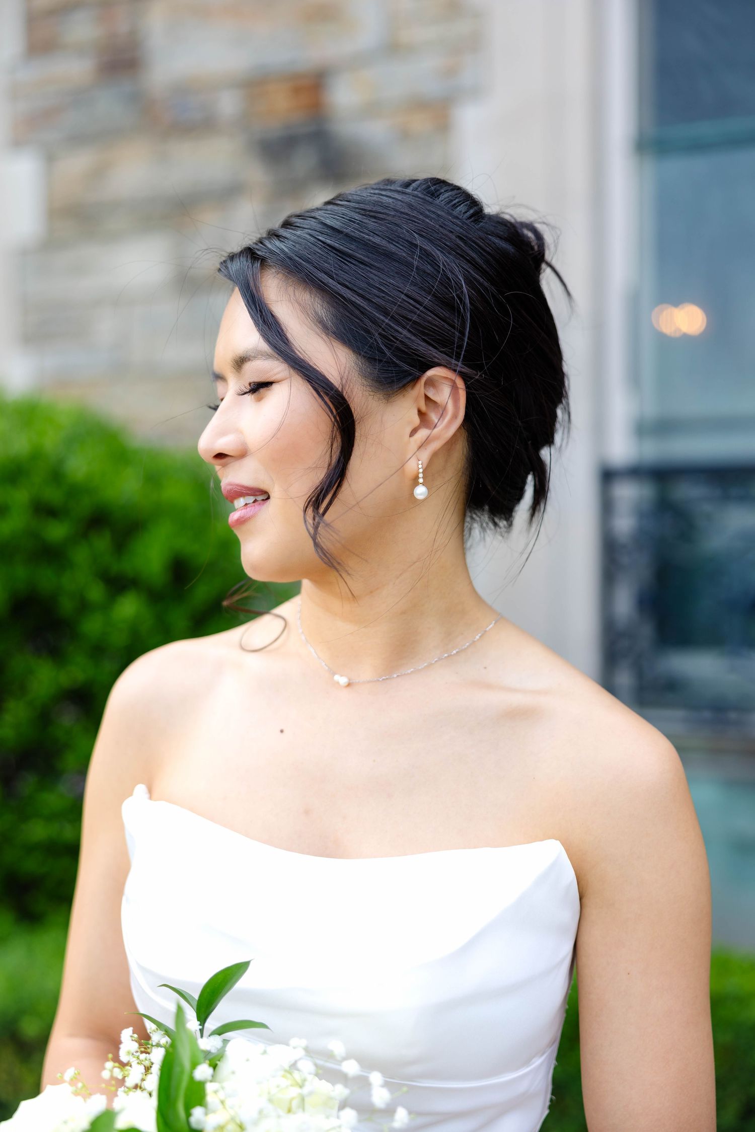Smiling Asian bride in strapless white gown with pearl earrings and necklace holding lily of the valley bouquet.