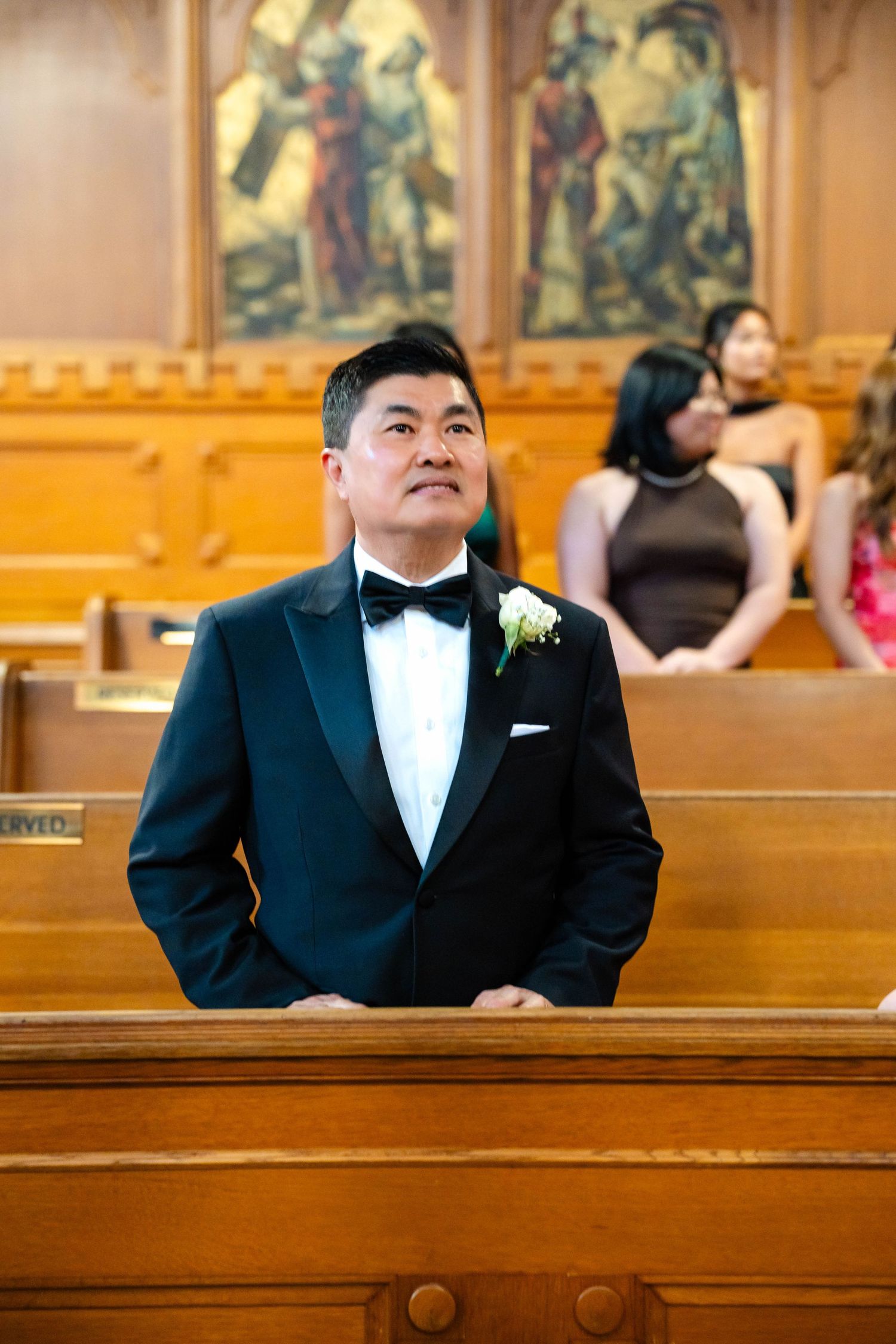 Groom in black tuxedo with boutonniere standing at church altar looking up during wedding ceremony.