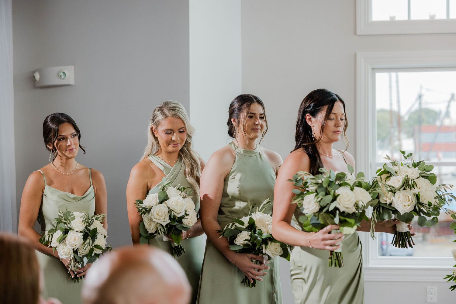 Four bridesmaids in sage green satin dresses holding white rose bouquets at wedding ceremony.