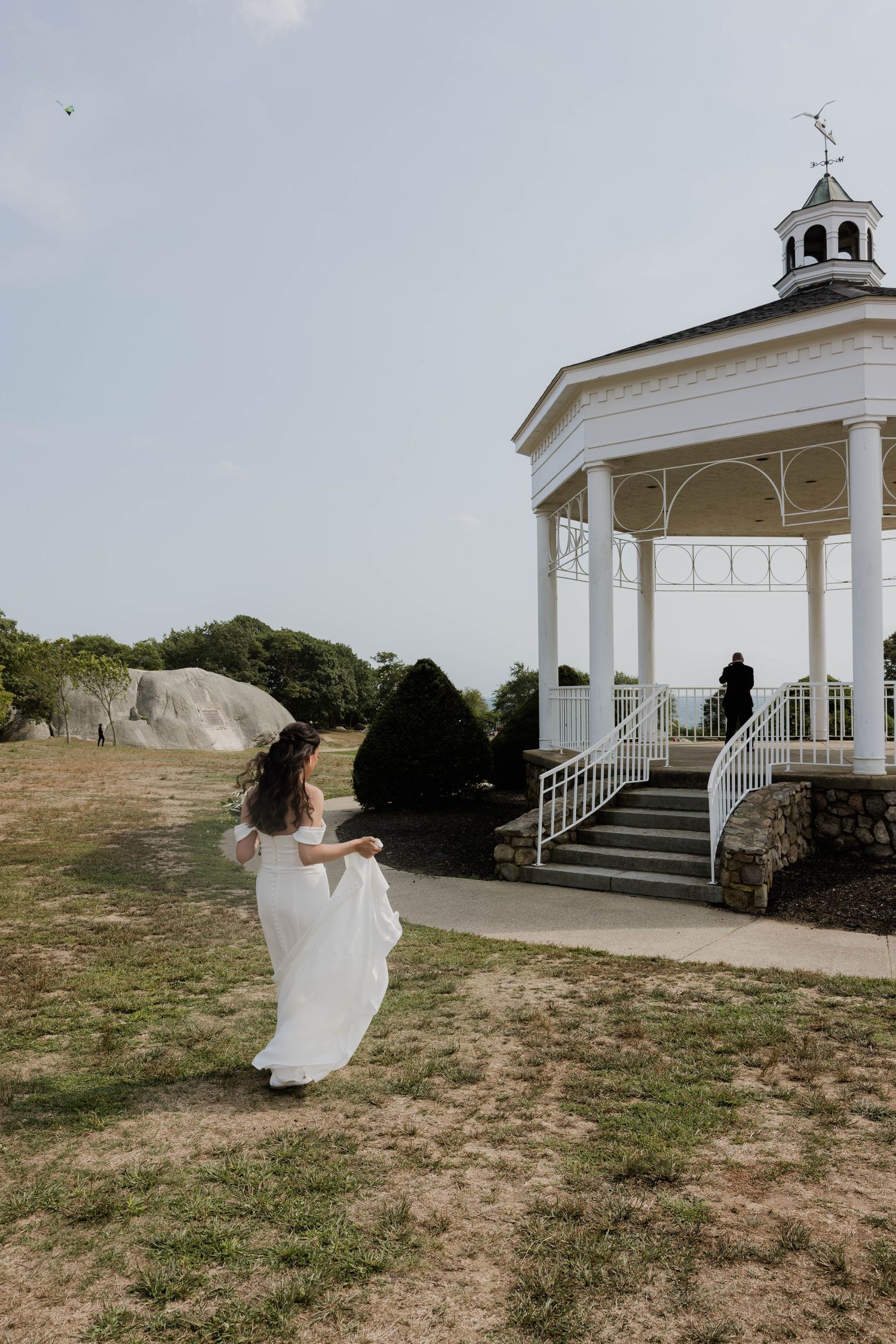 Bride in white gown walking toward a white gazebo on a sunny wedding day.