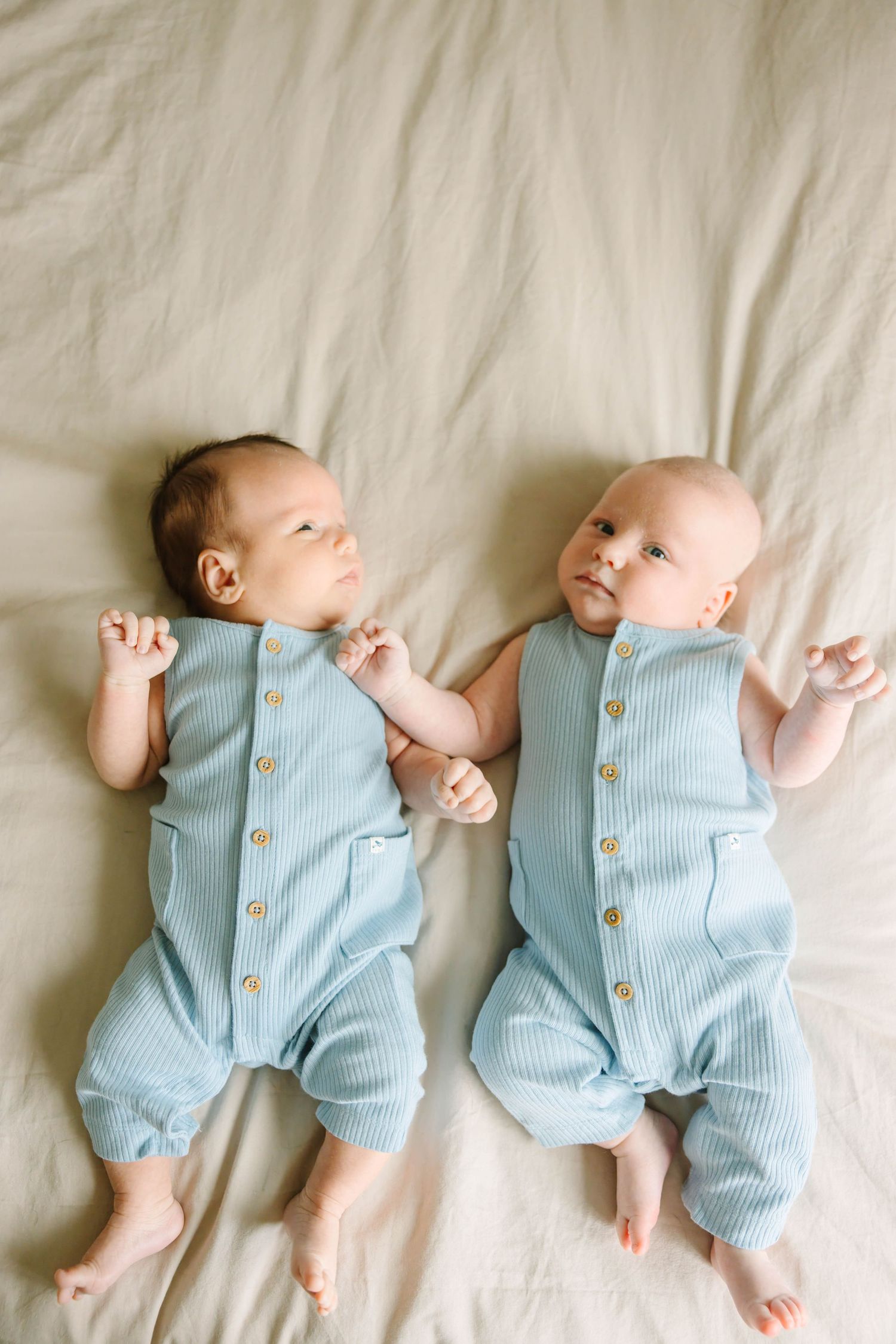 Twin newborn babies in matching blue ribbed button rompers lying side by side on a beige blanket.