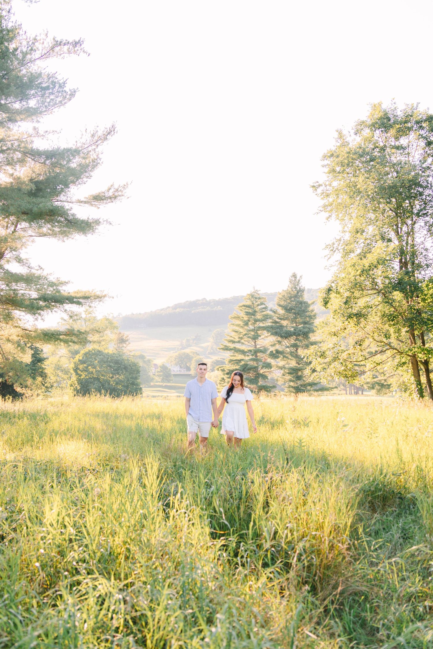 Couple holding hands in a sunlit meadow surrounded by lush green trees and rolling hills.