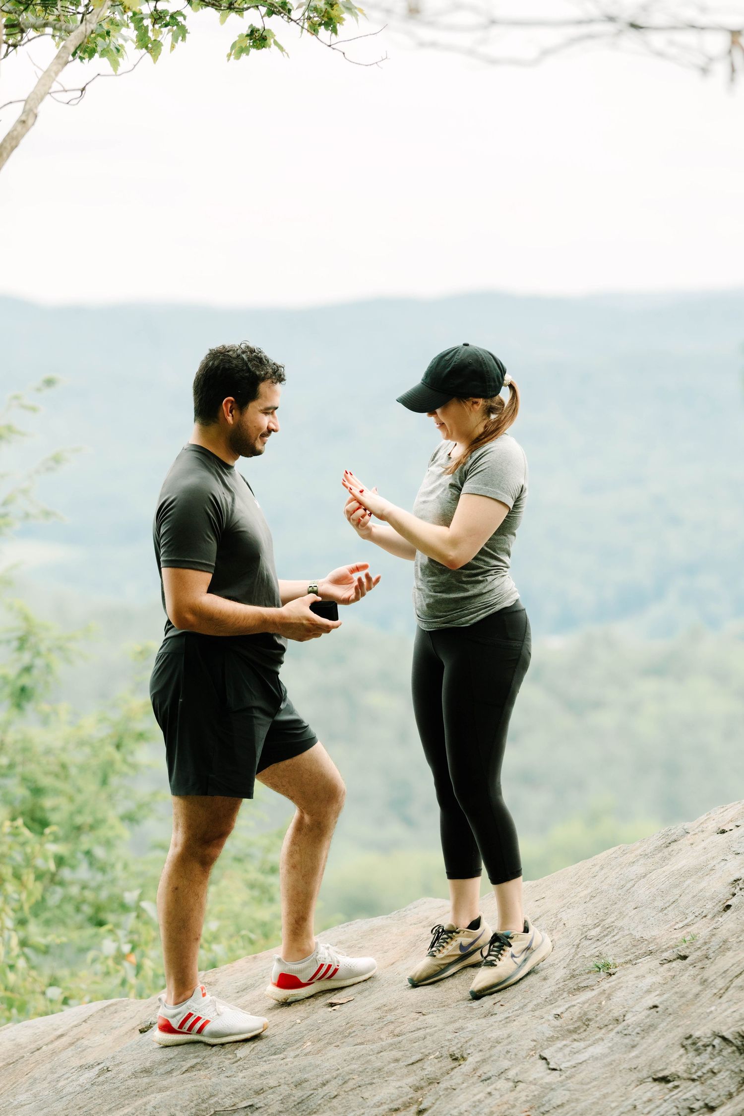 A man proposes to a woman on a rocky overlook with scenic mountain views in the background.