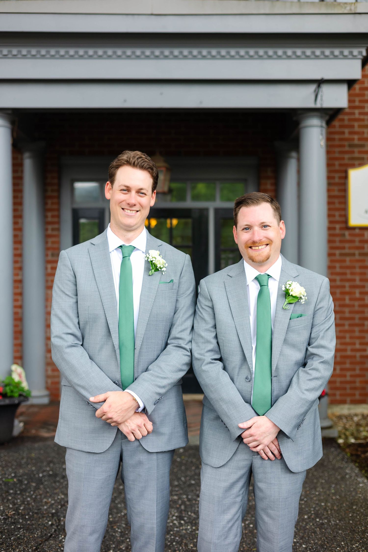 Two groomsmen in gray suits with green ties and boutonnieres smiling outside brick building entrance.