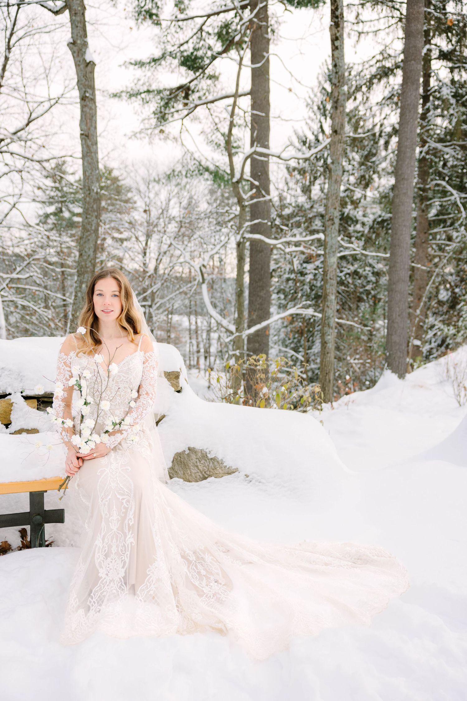 Smiling bride in lace wedding gown sitting on snowy bench surrounded by winter forest.