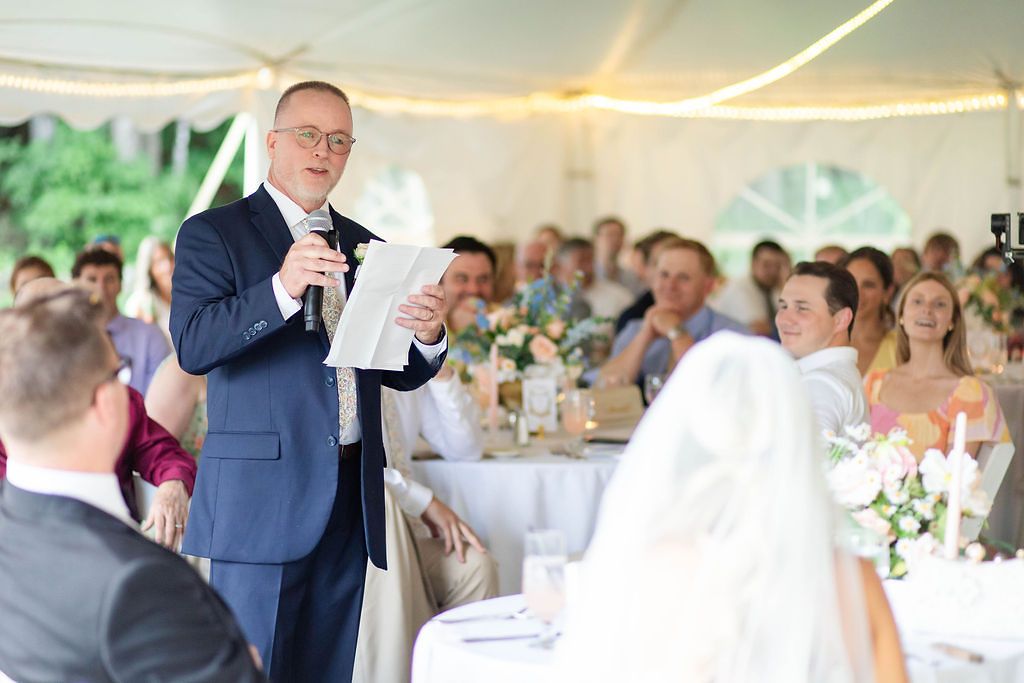Man in navy suit delivers a speech at a wedding reception held under a white tent.