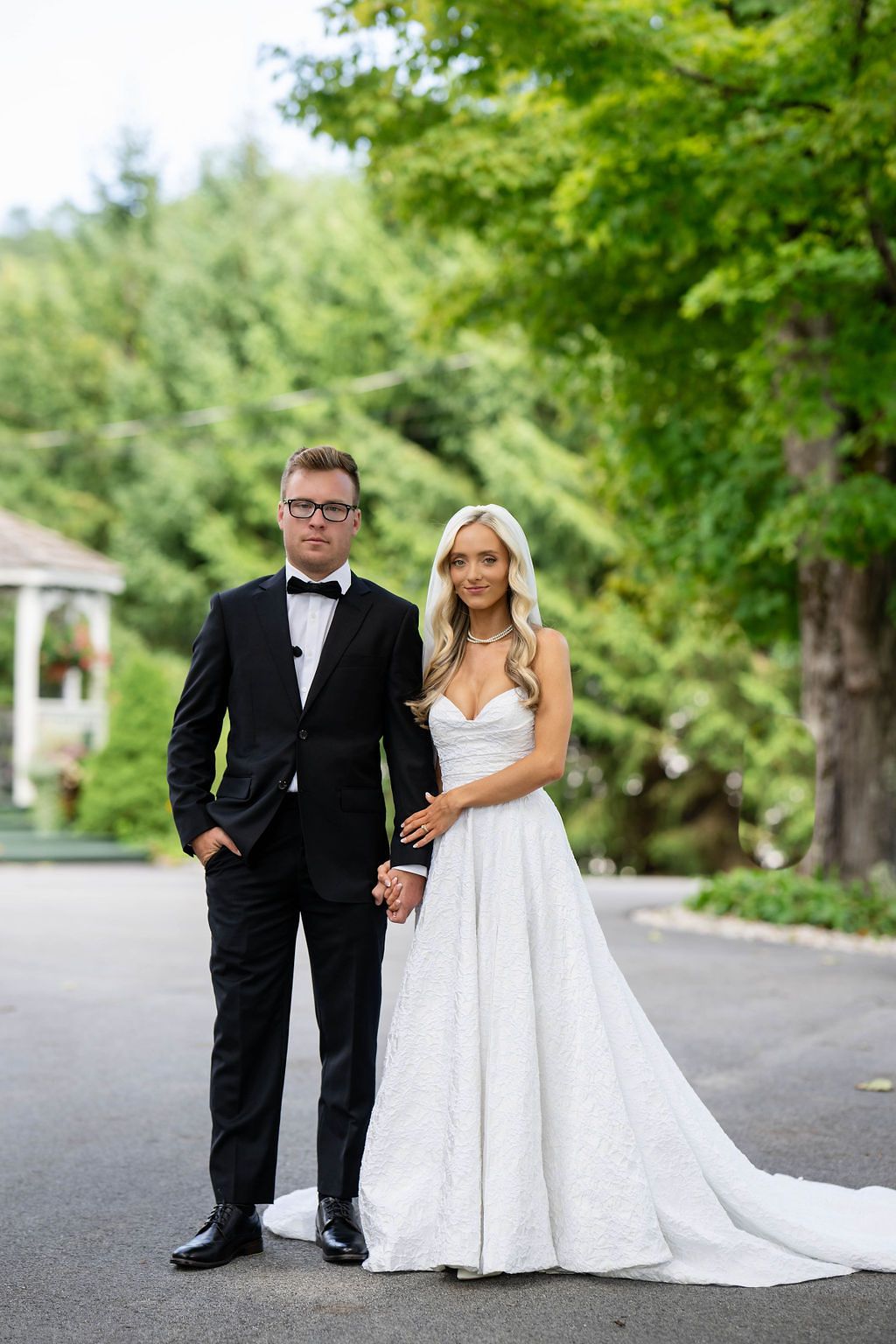 Elegant bride and groom posing in black tuxedo and strapless lace gown outdoors.