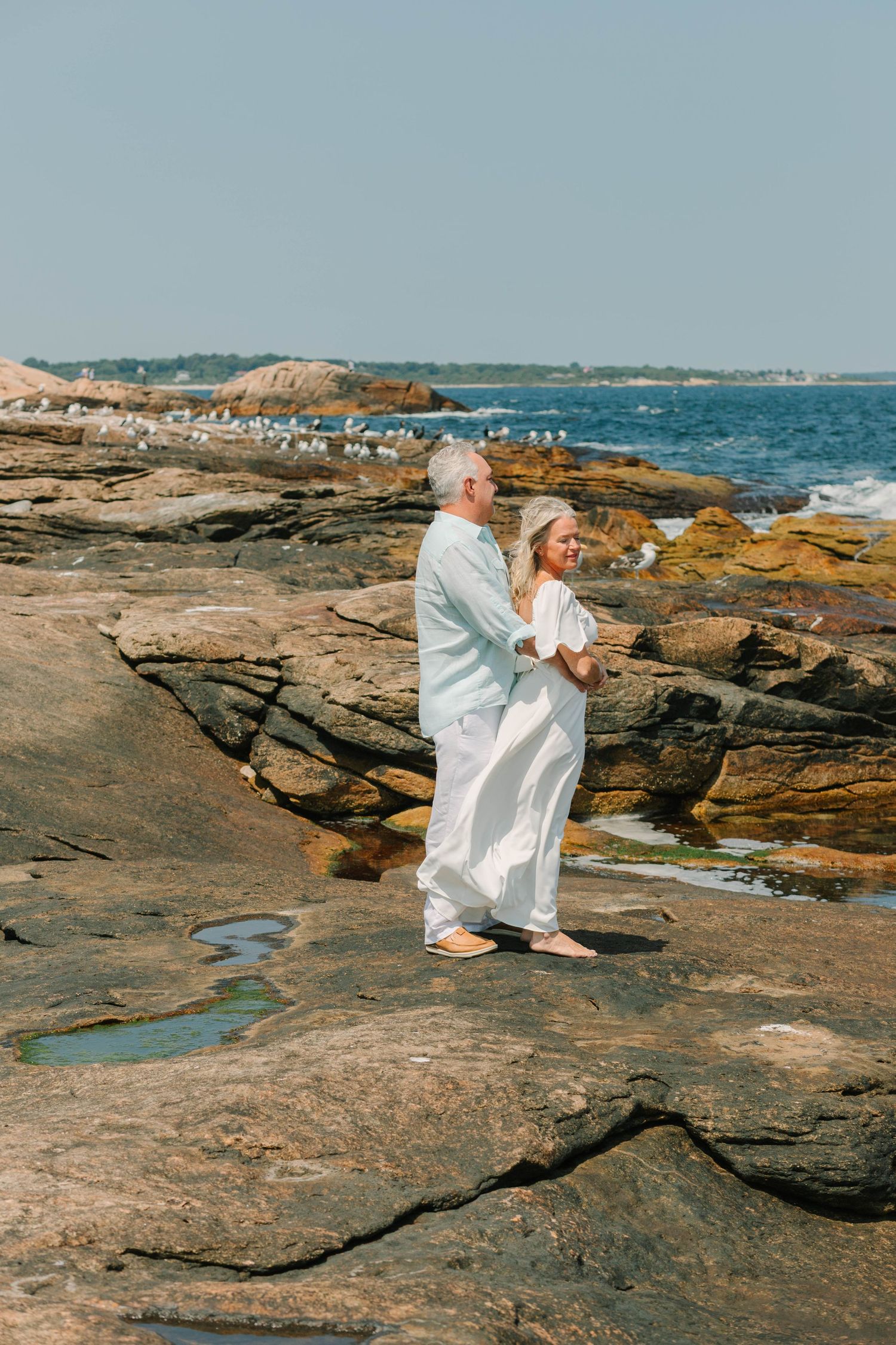 Couple in white attire standing on rocky coastal shore with ocean waves and seagulls in background.