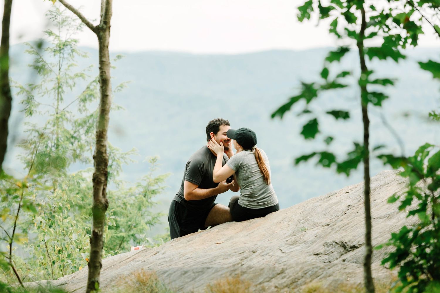 Couple sharing a kiss on a rocky overlook surrounded by lush green trees and misty mountain views.