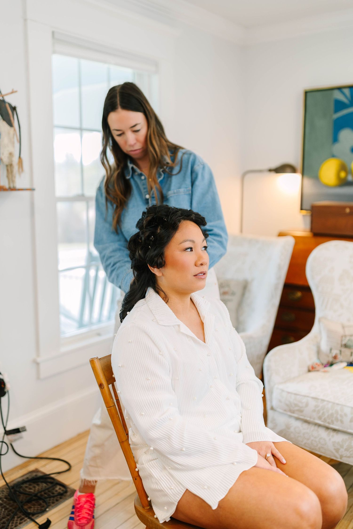 A hairstylist styling a seated Asian woman's dark braided updo in a bright white room.