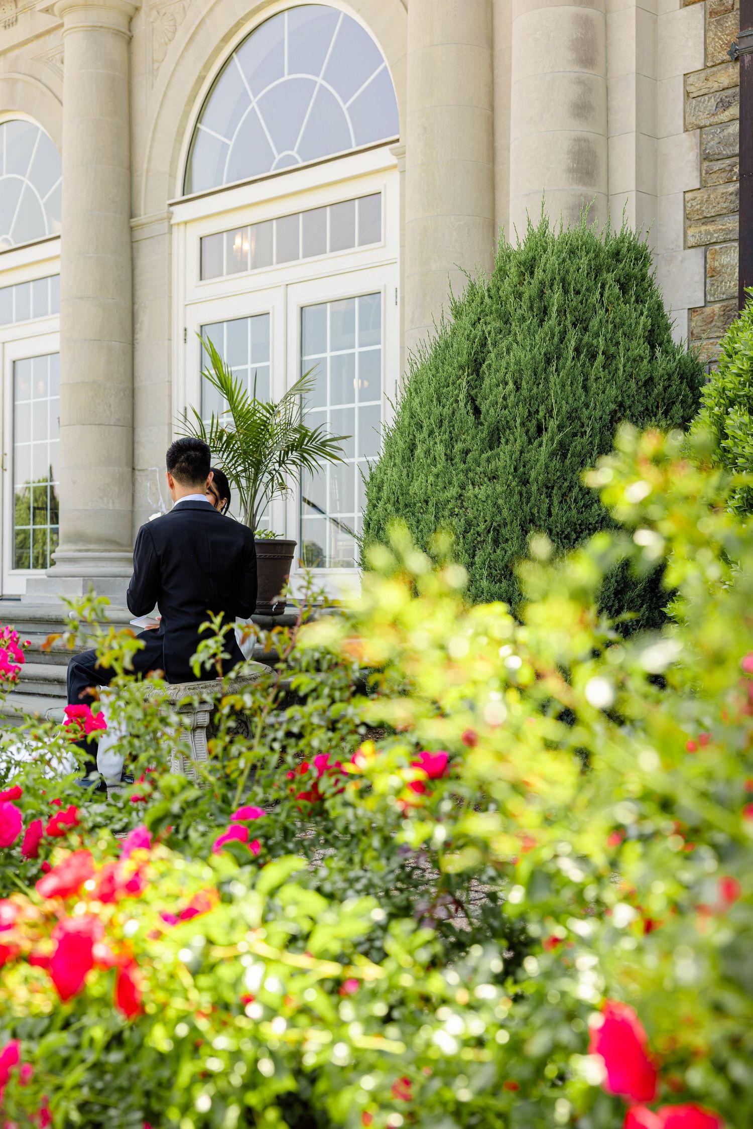 A man in a suit sits on steps of an elegant estate surrounded by lush gardens with colorful flowers.
