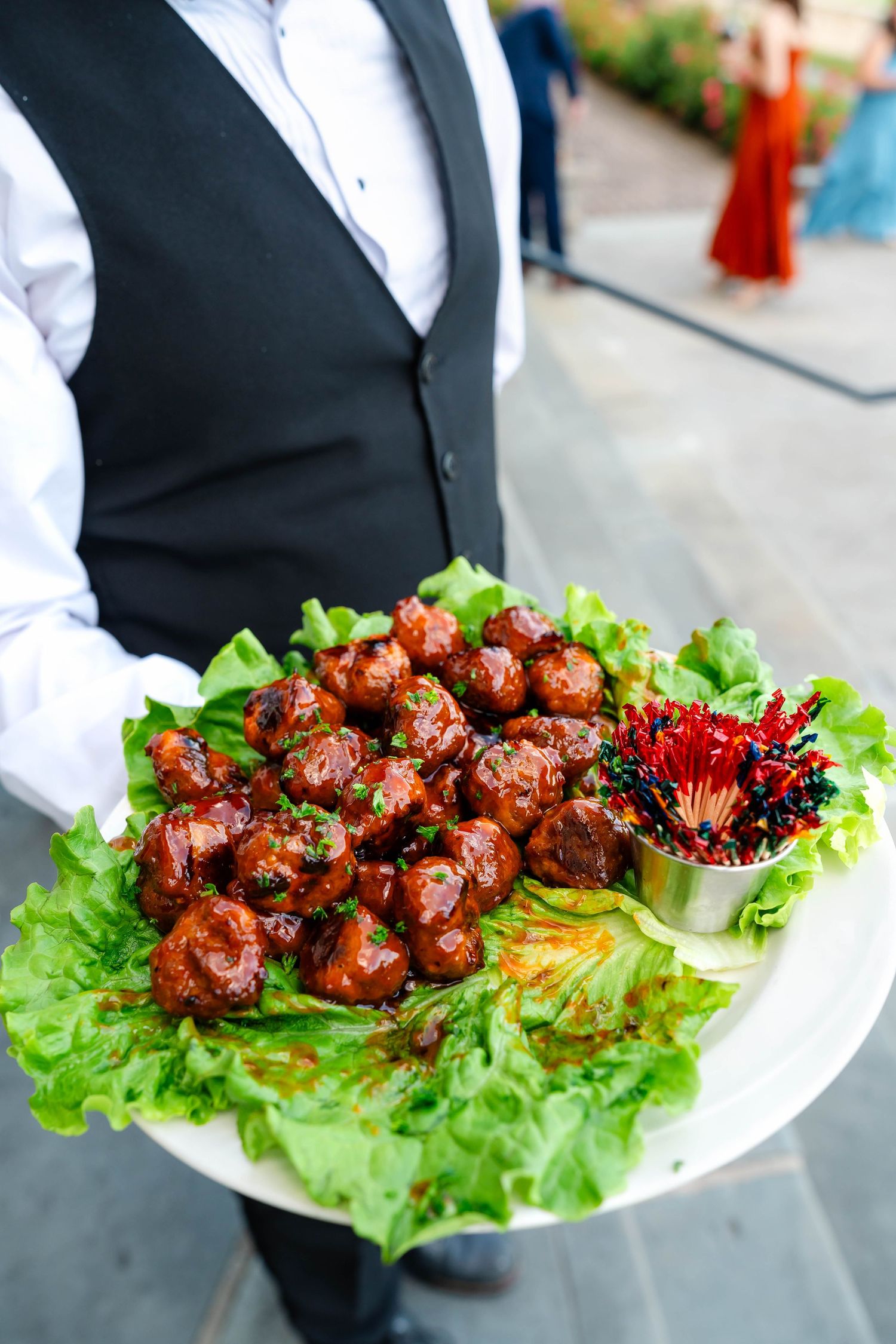 Waiter holds a large platter of glazed meatballs on lettuce with decorative cocktail picks.