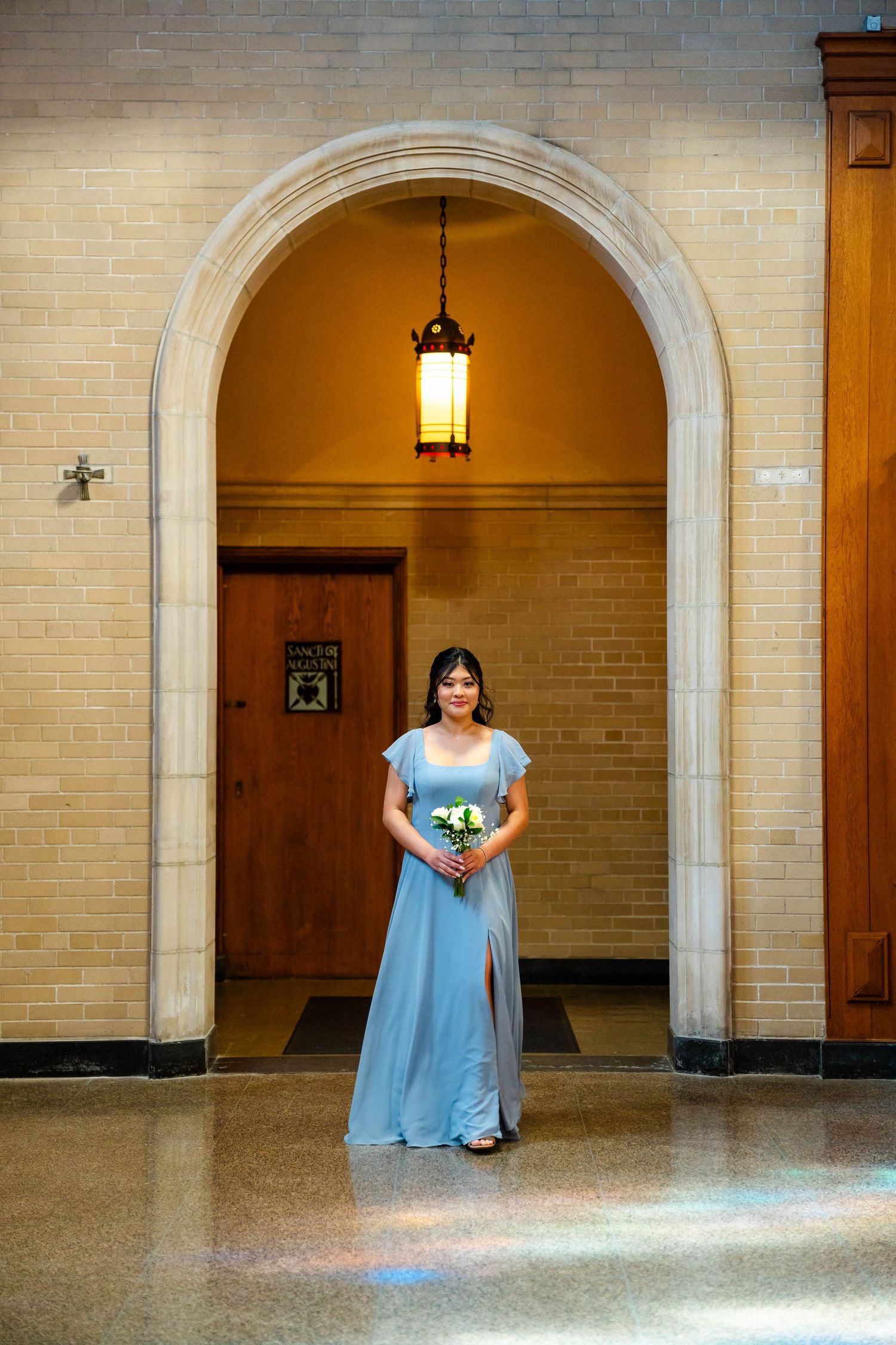 Bridesmaid in blue gown holding bouquet standing under stone archway with vintage lantern.