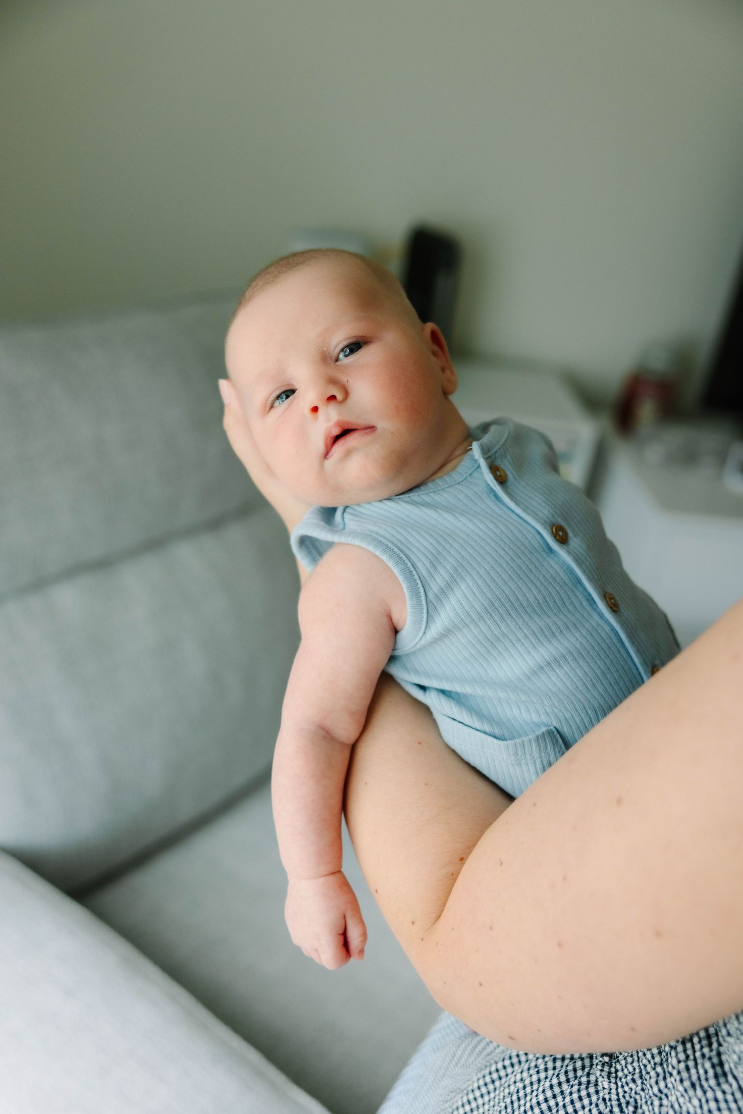 A newborn baby in a blue ribbed onesie being held by a parent on a gray sofa.