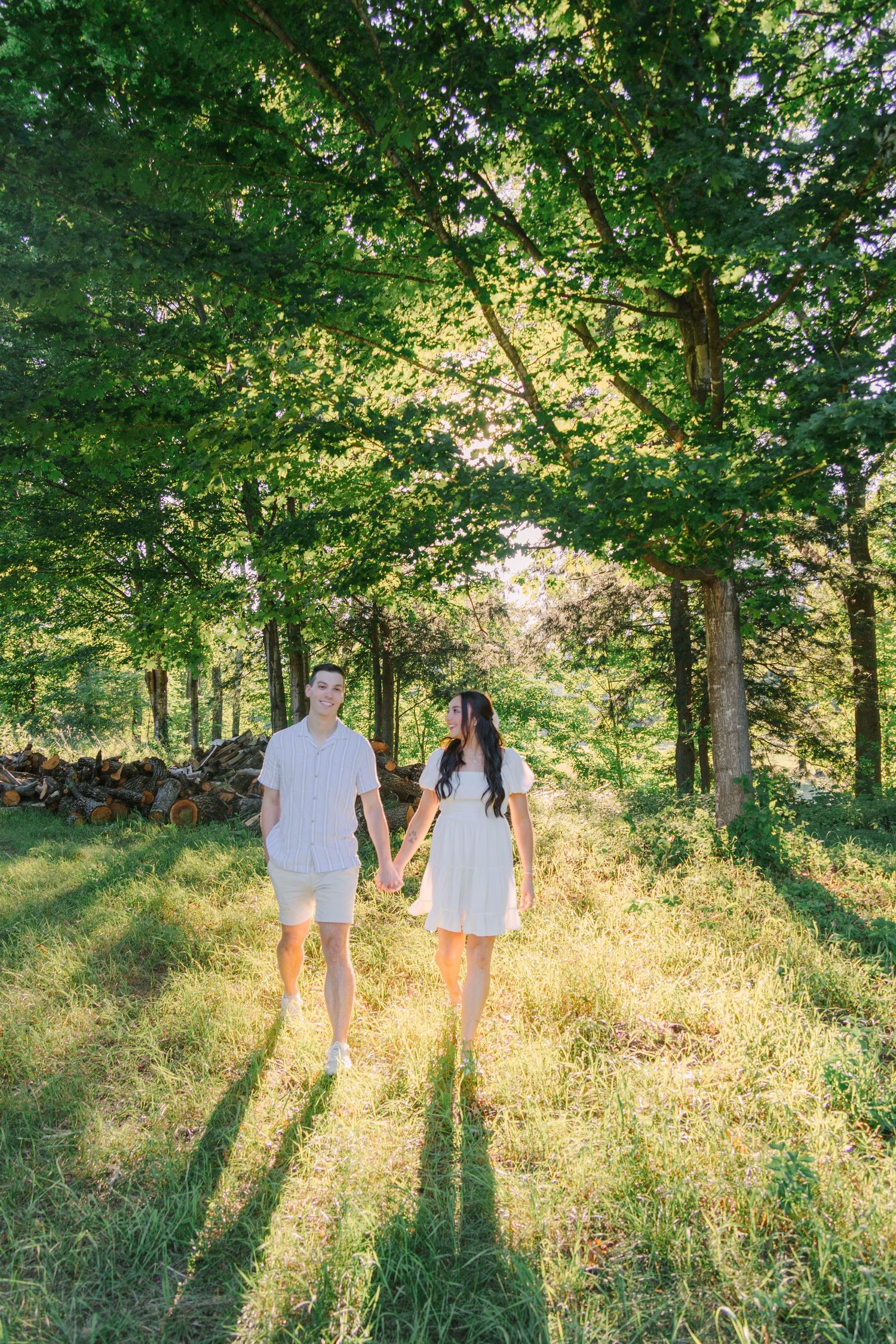 Couple in white outfits holding hands while walking through a sunlit green forest path on a sunny day.