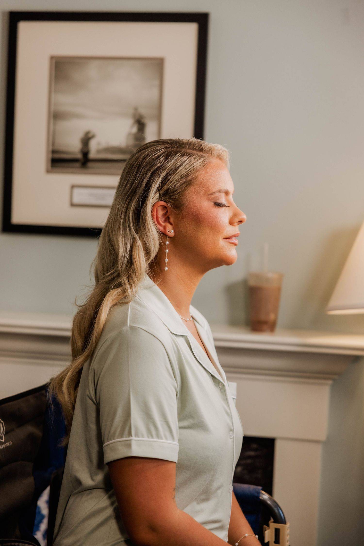 Blonde woman in sage green top sits with eyes closed, wearing pearl drop earrings in an elegant room.