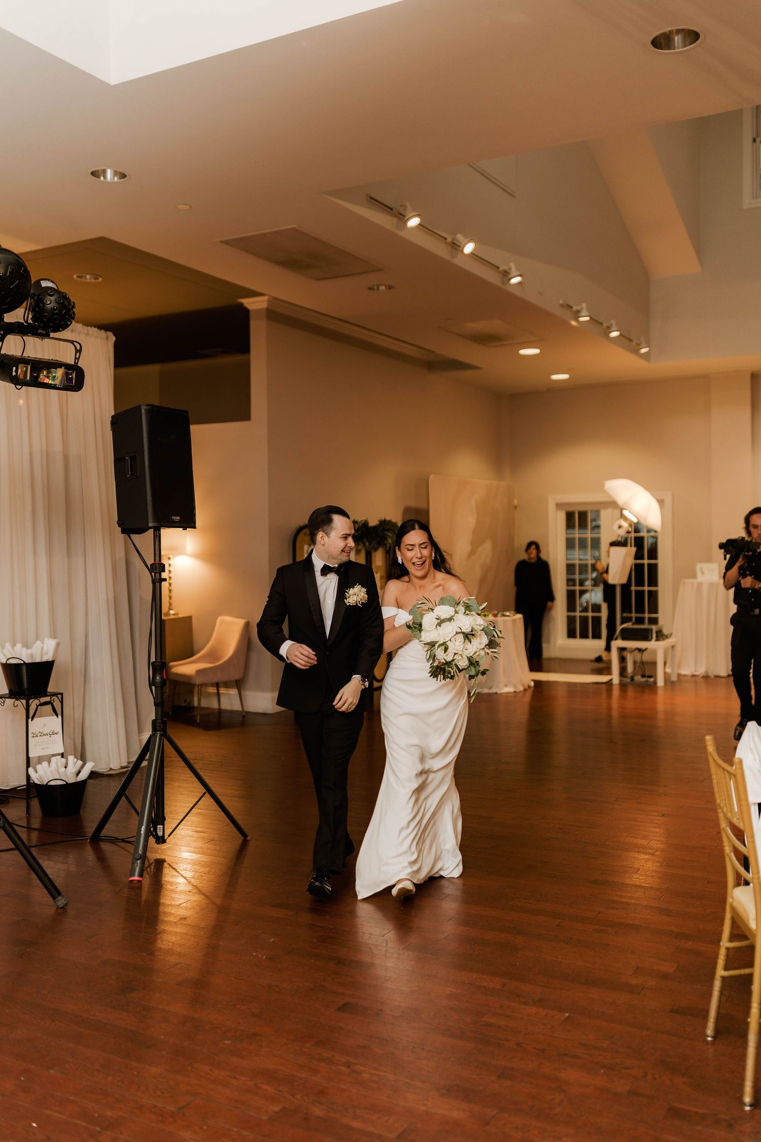 Bride and groom walk hand-in-hand into their elegant wedding reception venue with warm lighting and hardwood floors.