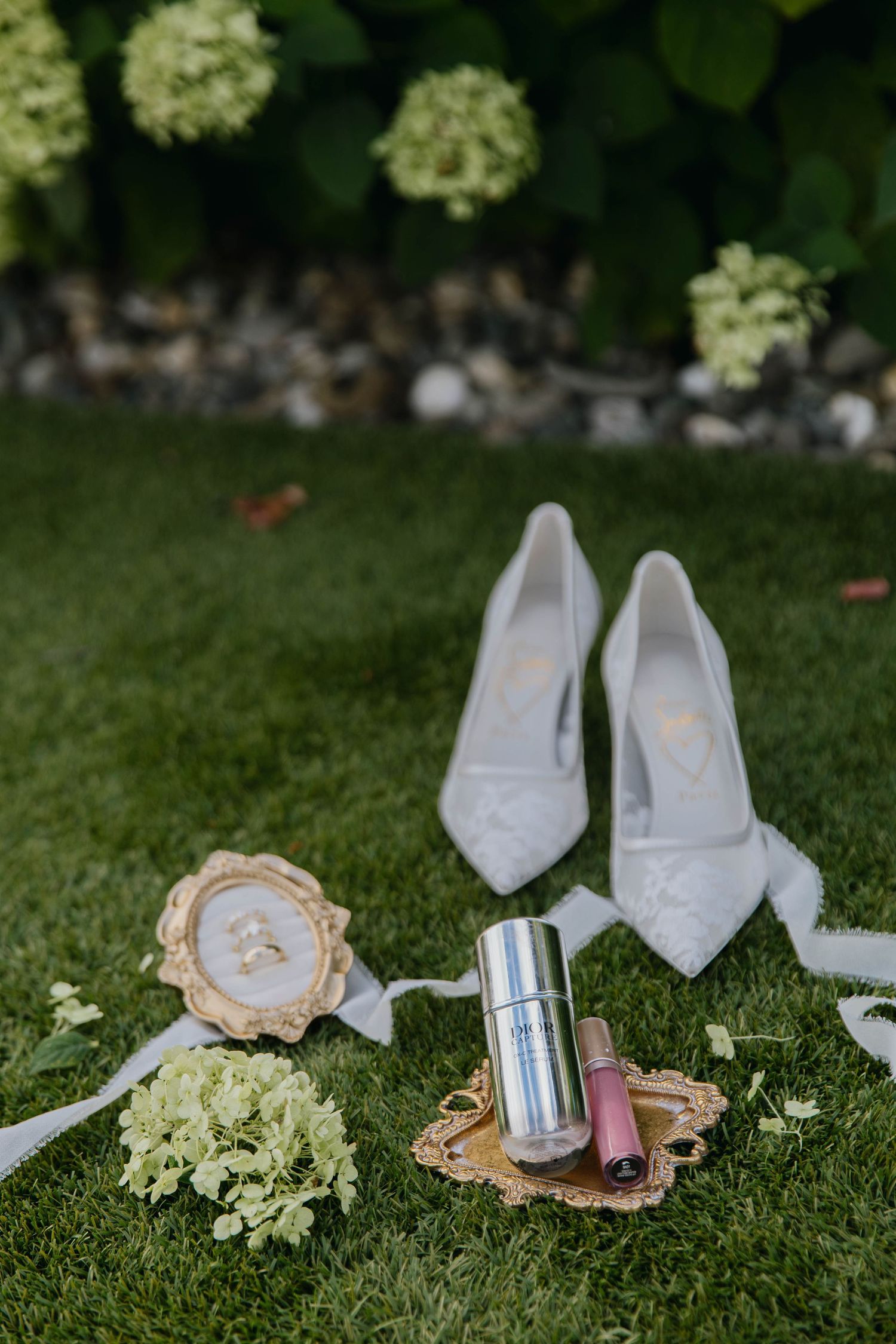 Bridal flat lay on green grass featuring white lace heels, wedding rings, lipstick, and hydrangea flowers.