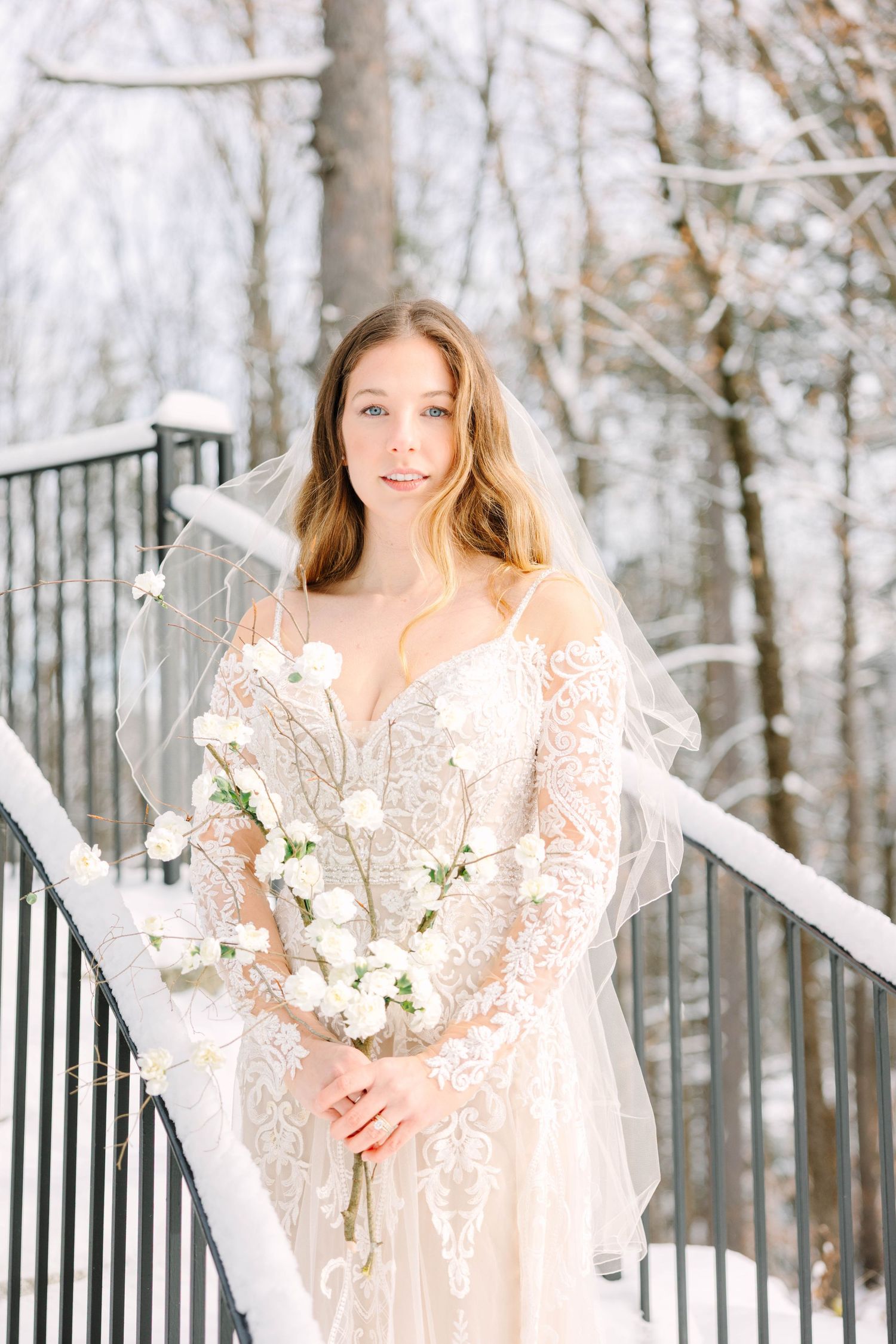 Close-up of bride in lace wedding dress holding white floral bouquet on snow-covered railing in winter forest.