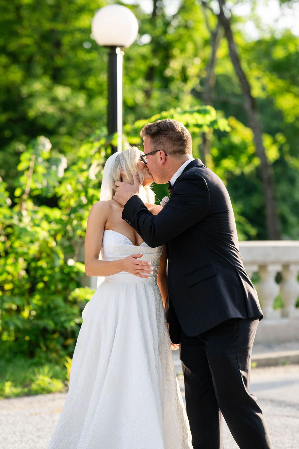 Bride and groom share a romantic kiss outdoors surrounded by lush green trees on their wedding day.