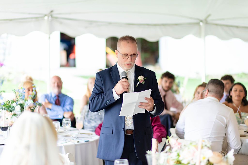 Man in navy suit giving speech at wedding reception under white tent with guests seated.