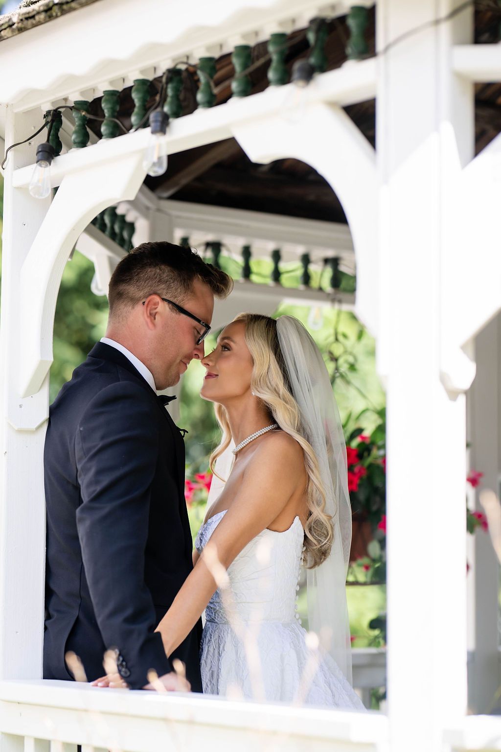 Bride and groom share a romantic moment in a white gazebo, surrounded by lush greenery and flowers on their wedding day.