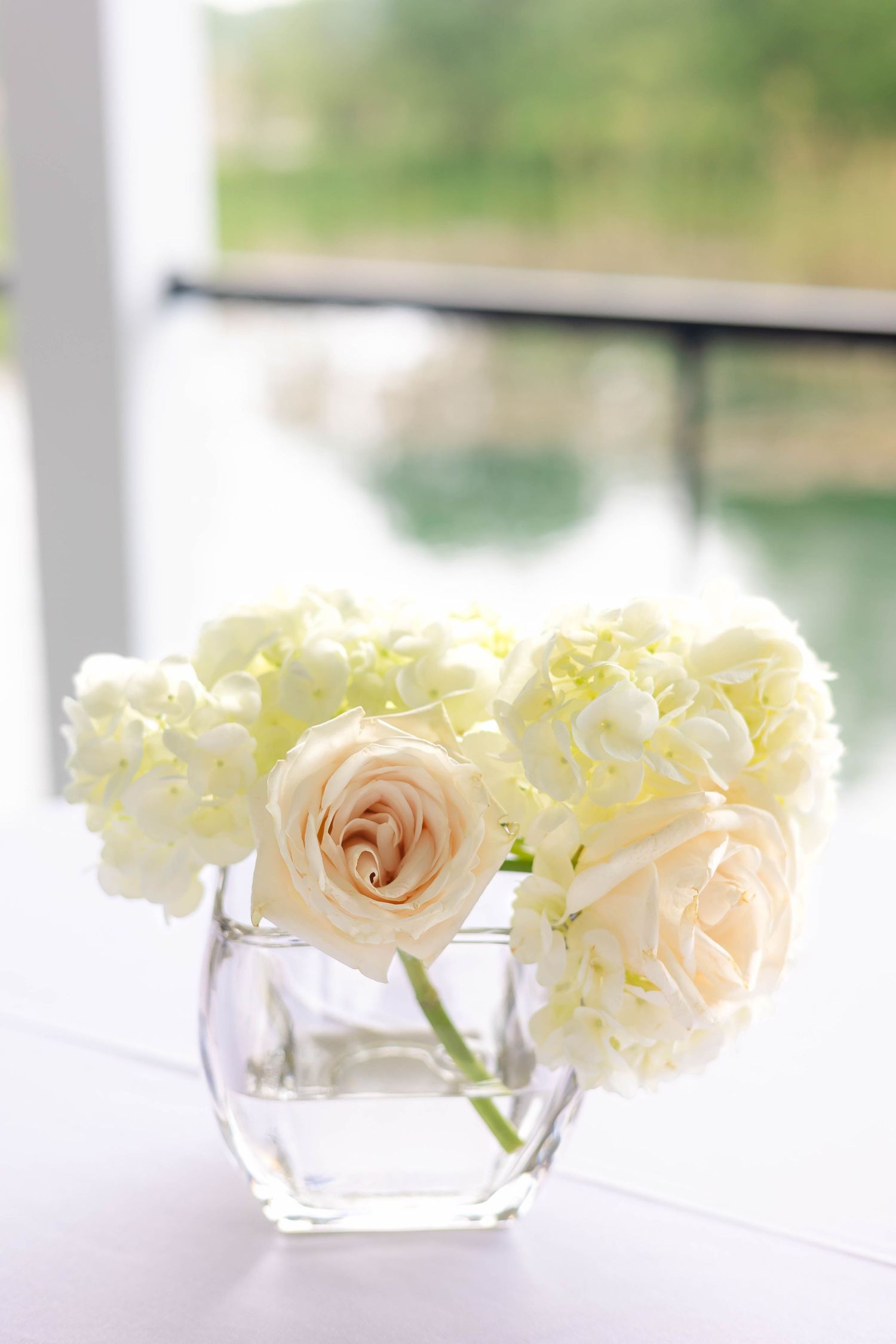 White roses and hydrangeas in clear glass vase on white table near window with garden view.