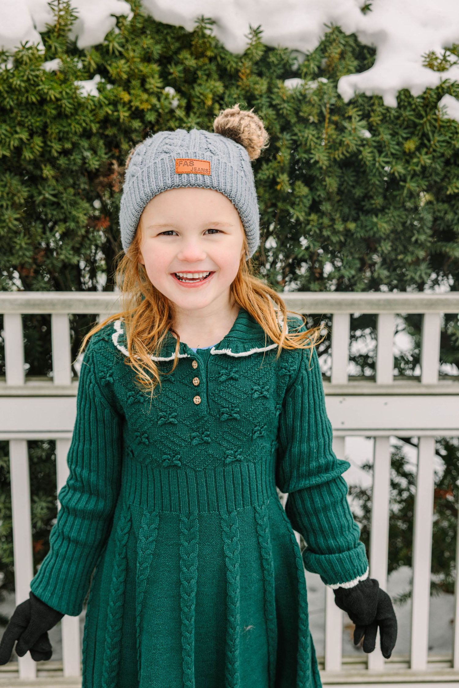 A cheerful young girl in a green knit dress and gray pom-pom beanie smiles outdoors in a snowy winter setting.
