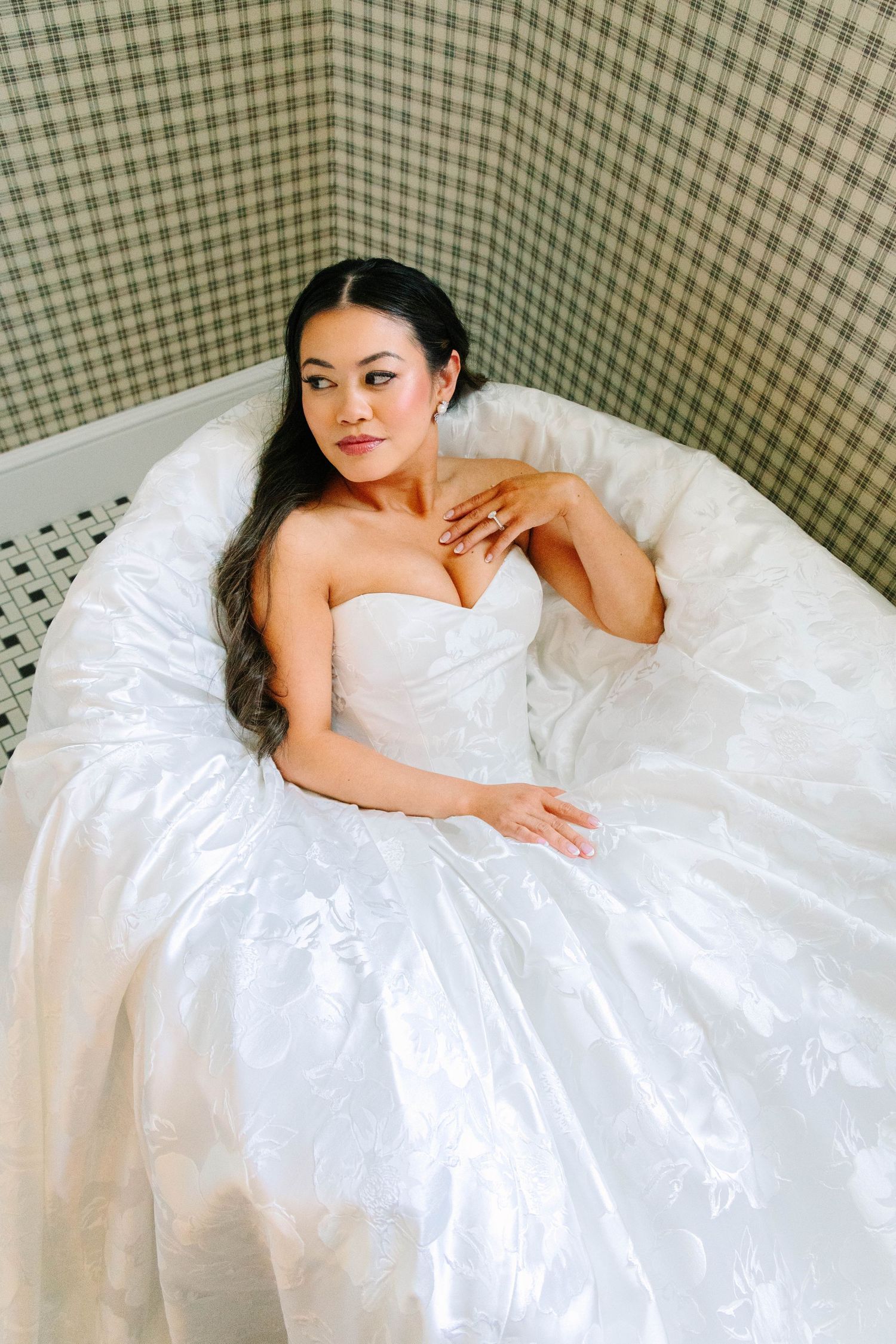 Elegant bride in a strapless ballgown poses gracefully in a clawfoot bathtub with plaid wallpaper backdrop.