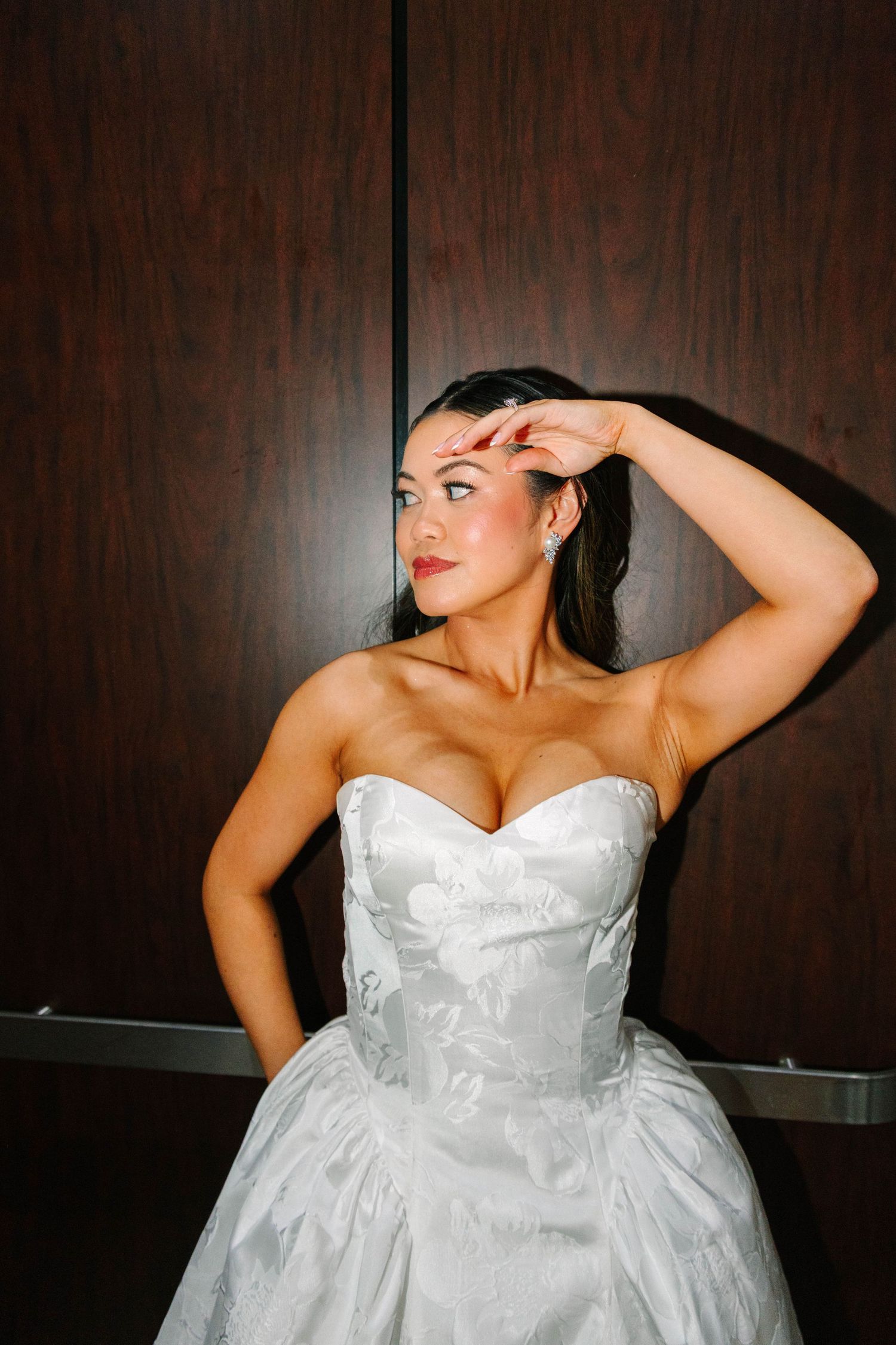 Bride in strapless white floral ballgown posing dramatically against dark wood paneled wall.