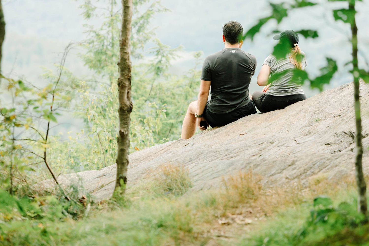 Couple sitting on a large rock in a lush forest, enjoying nature views while one person holds a smartphone.